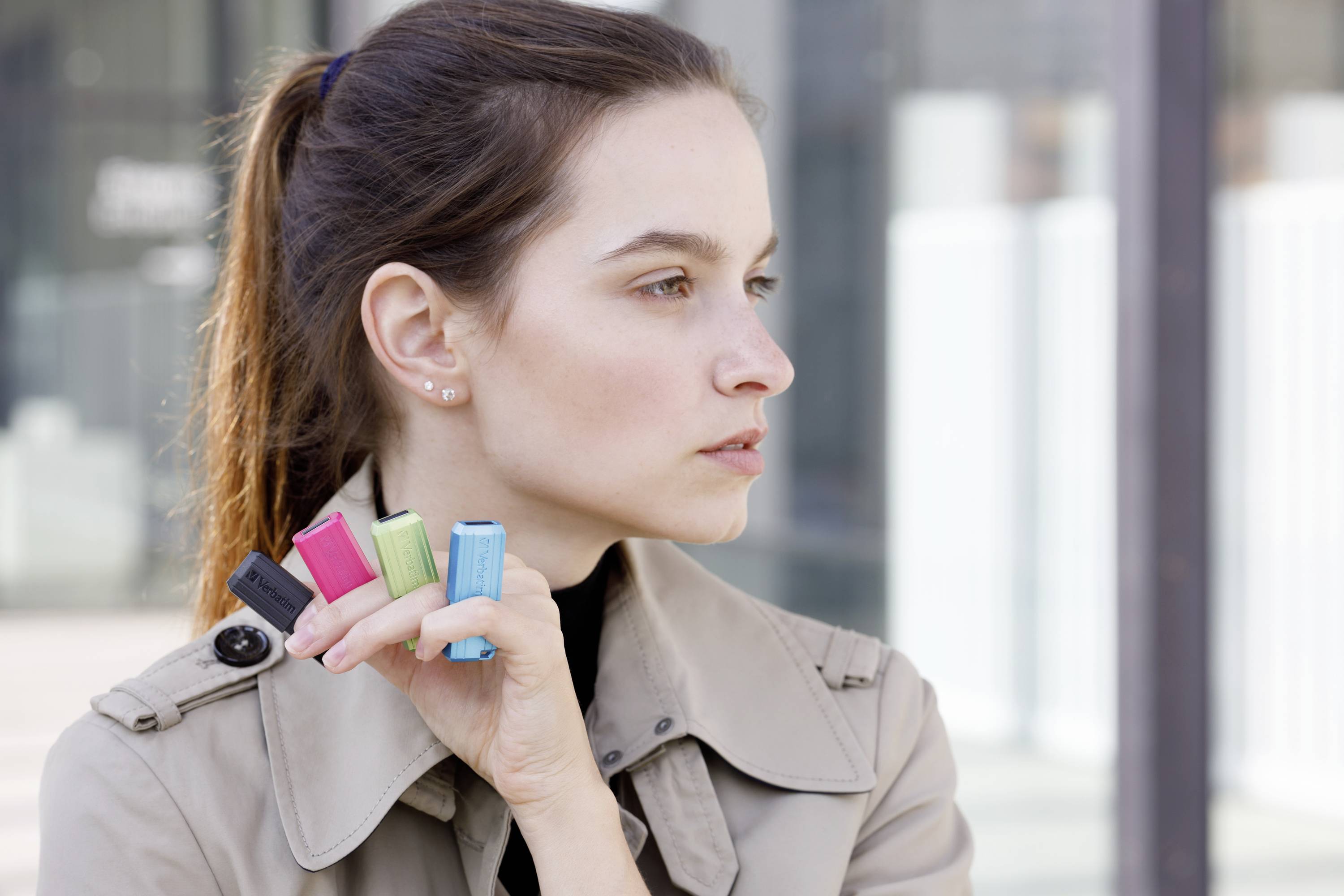 Eine Person im Profil trägt vier bunte Lippenstiftetuis an den Fingern und schaut konzentriert in die Ferne, im Freien.