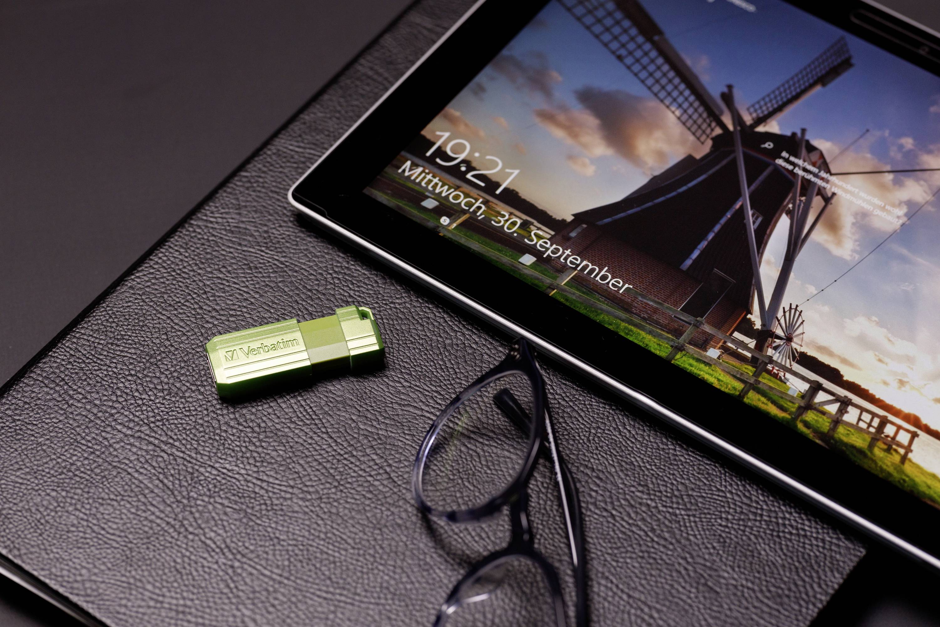 Ein grüner USB-Stick neben einer Brille auf einem schwarzen Untergrund. Im Hintergrund zeigt ein Bildschirm eine Windmühle und Datum/Zeit.