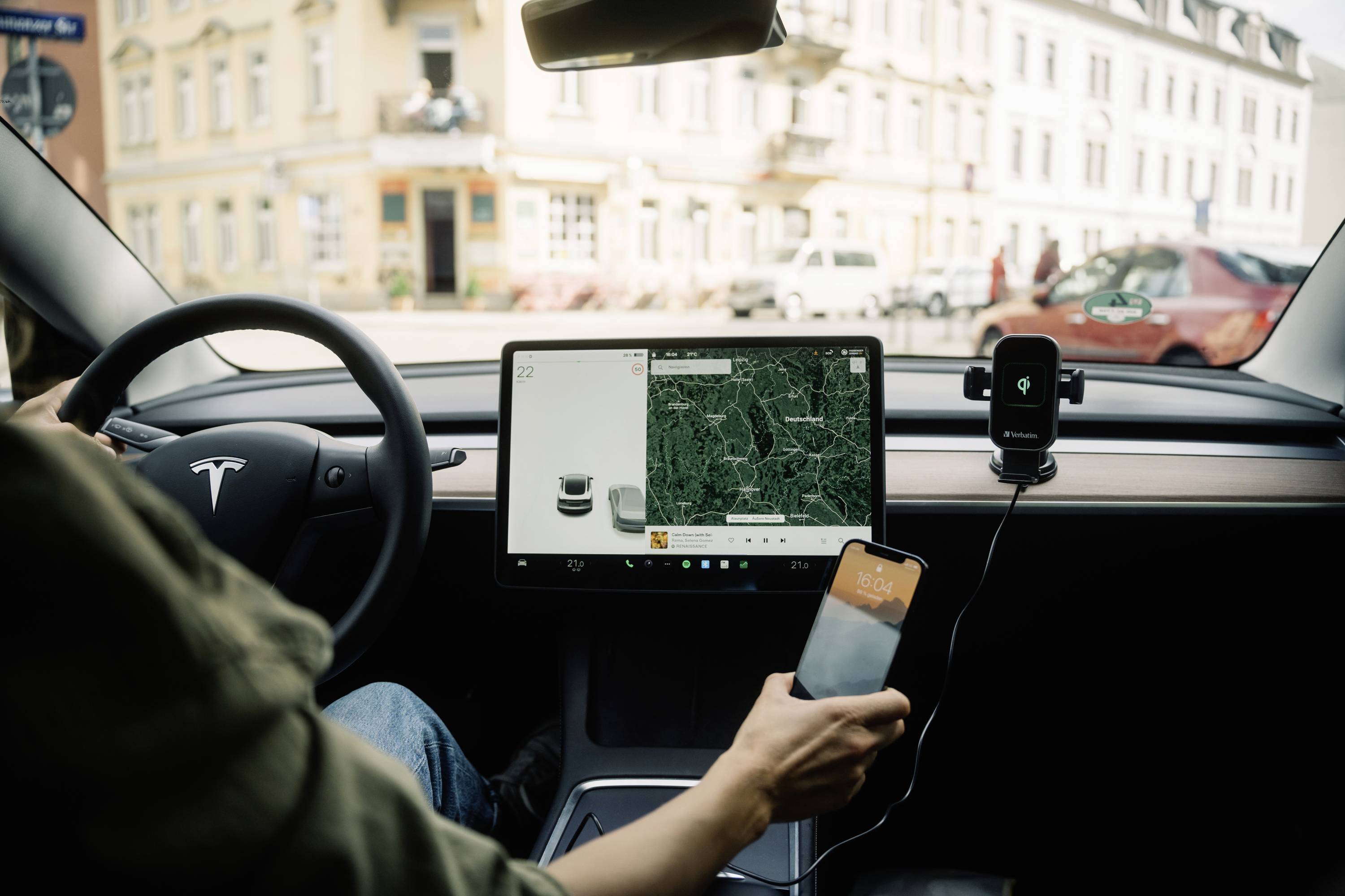 Person fährt Auto mit großem Touchscreen-Display, zeigt Kartenanwendung. Außenansicht einer Stadt mit Gebäuden und geparkten Autos.
