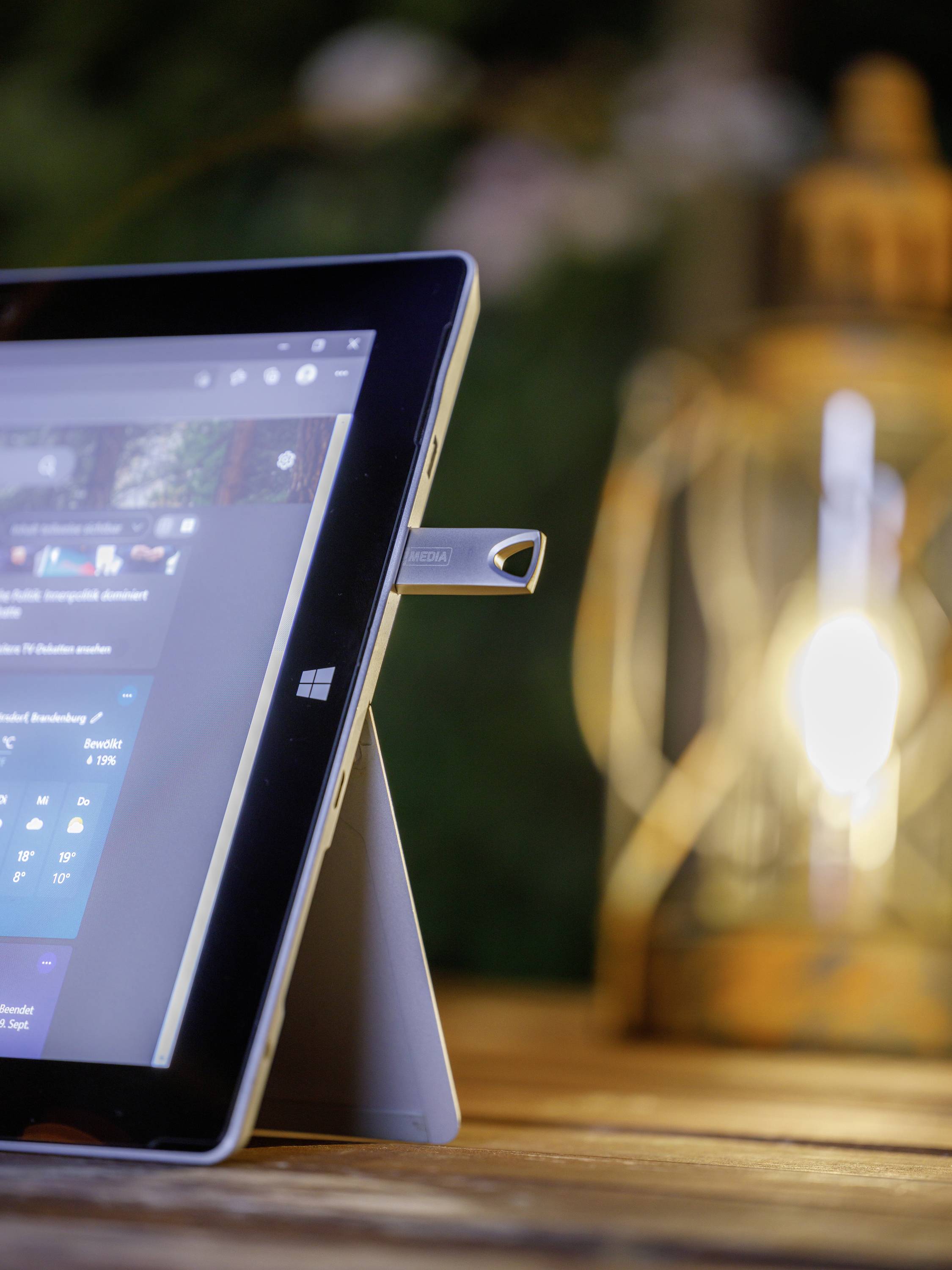 Ein Tablet steht auf einem Holztisch, mit einem USB-Stick eingesteckt. Im Hintergrund leuchtet eine Laterne warm.