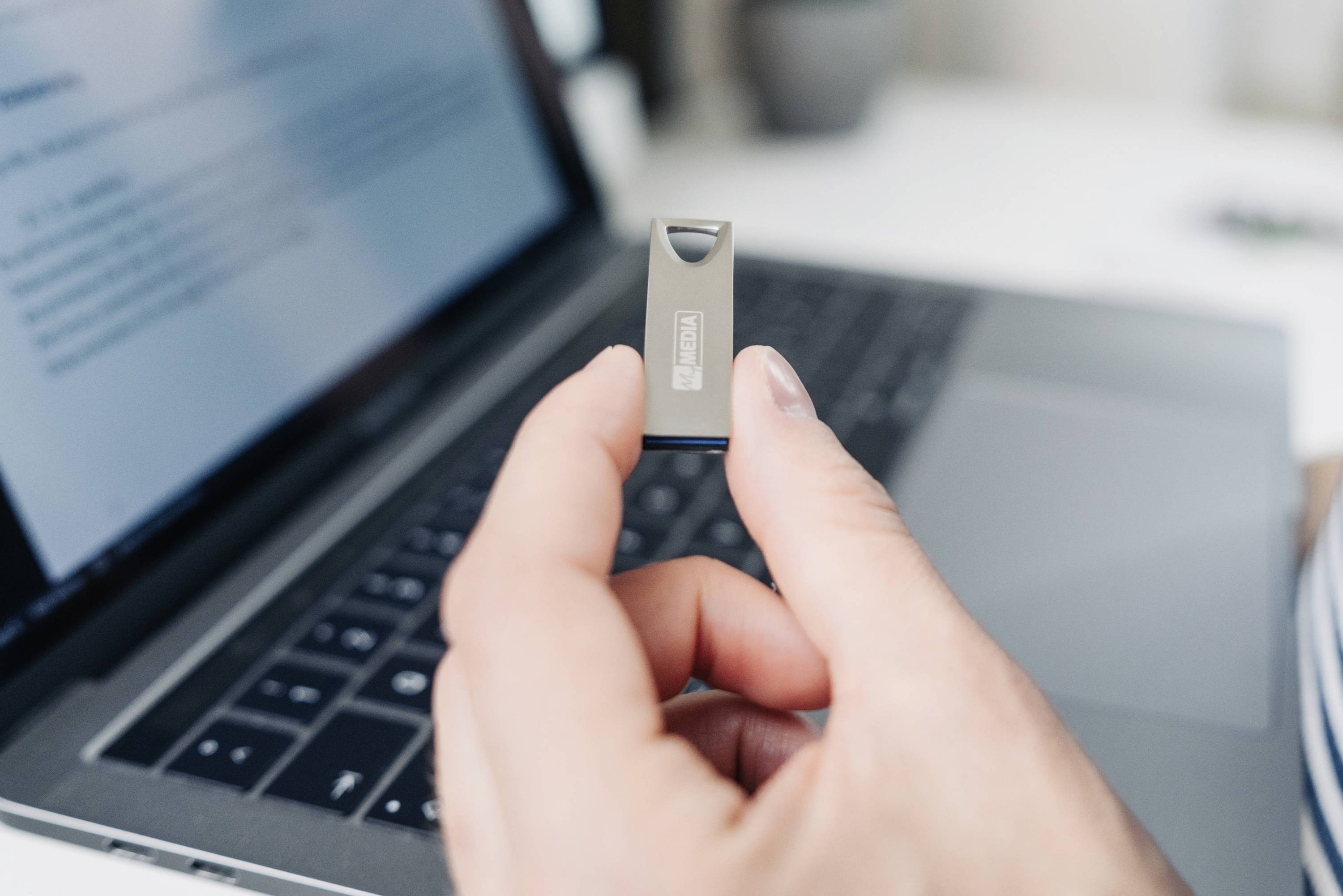 Person hält einen USB-Stick vor einem geöffneten Laptop-Bildschirm. Der Bildschirm zeigt unscharfen Text. Thema: Datenübertragung.