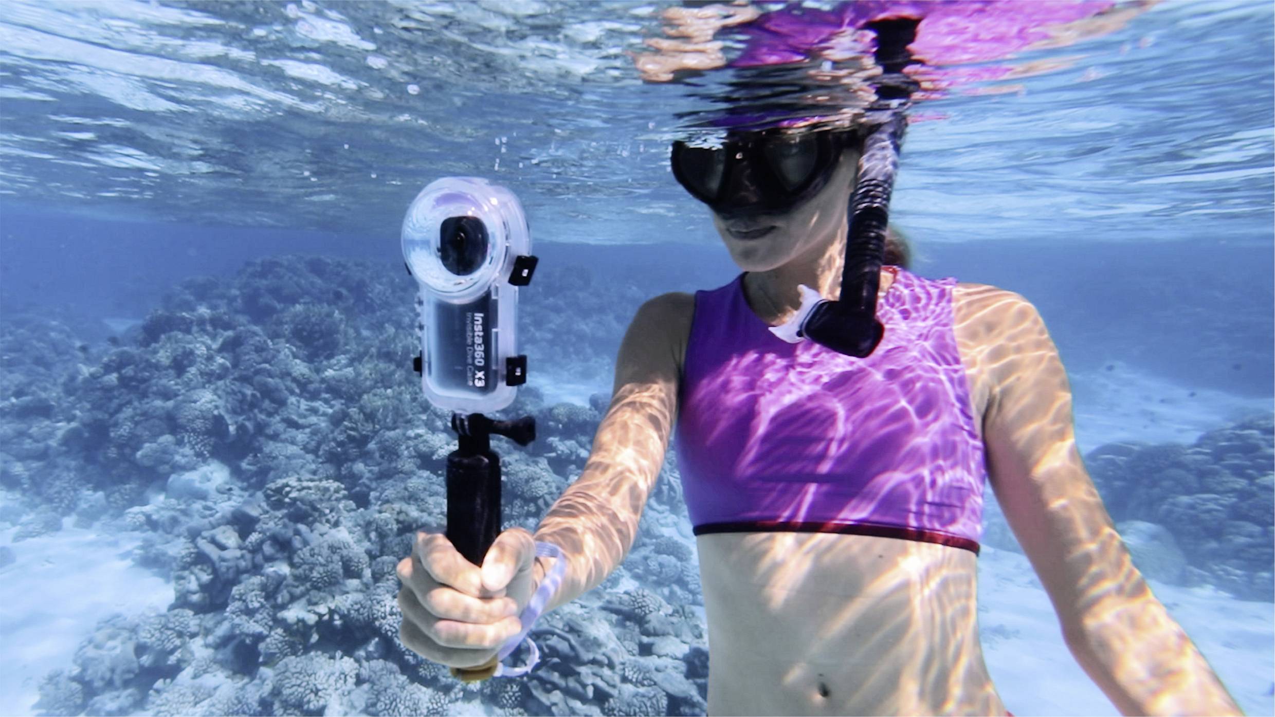 Eine Person im lilafarbenen Badeanzug schnorchelt unter Wasser und hält eine Kamera. Im Hintergrund sind Korallen zu sehen.