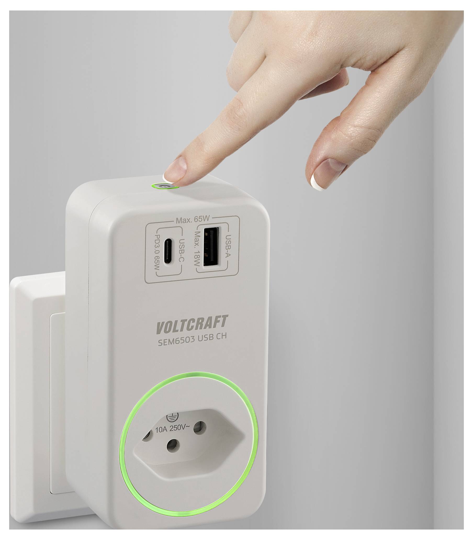 Eine Hand drückt einen Knopf auf einem Voltcraft USB-Ladegerät, das in eine Steckdose gesteckt ist, mit zwei USB-Anschlüssen und einer Steckdose.