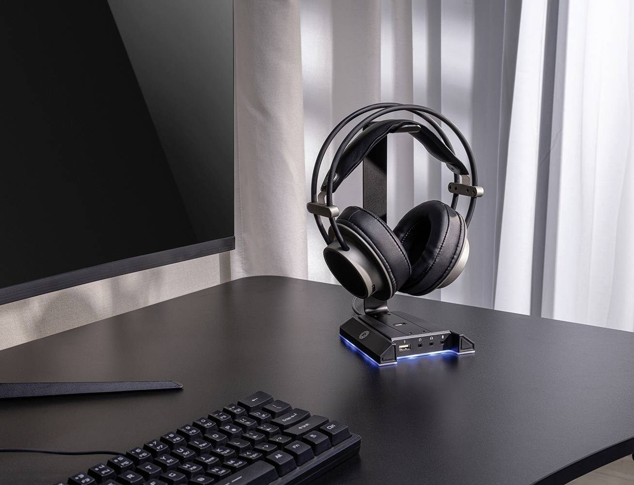 Ein Schreibtisch mit Monitor, Tastatur und einem Paar Kopfhörer auf einem Ständer. Sonnenlicht fällt durch den Vorhang.