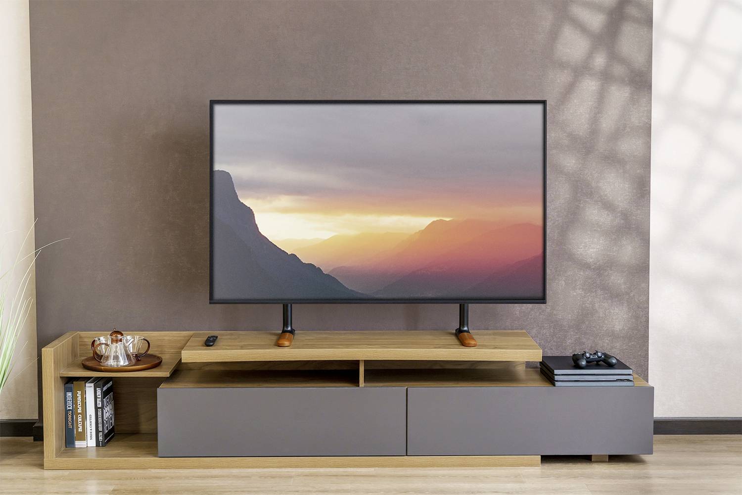 Flachbildfernseher auf Holzschrank vor brauner Wand, zeigt Sonnenuntergang in Berglandschaft. Links ein Tablett mit Tassen.