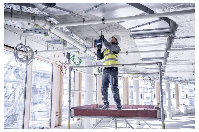 Ein Bauarbeiter in einer Sicherheitsweste und einem Schutzhelm verwendet eine Bohrmaschine, um Ausrüstung an der Decke eines modernen Gebäudes im Bau zu installieren.