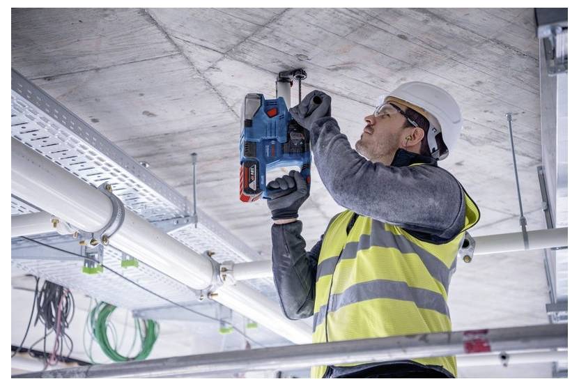 Ein Arbeiter in einer Warnweste und mit Helm verwendet ein Elektrowerkzeug, um in eine Betondecke auf einer Baustelle zu bohren.