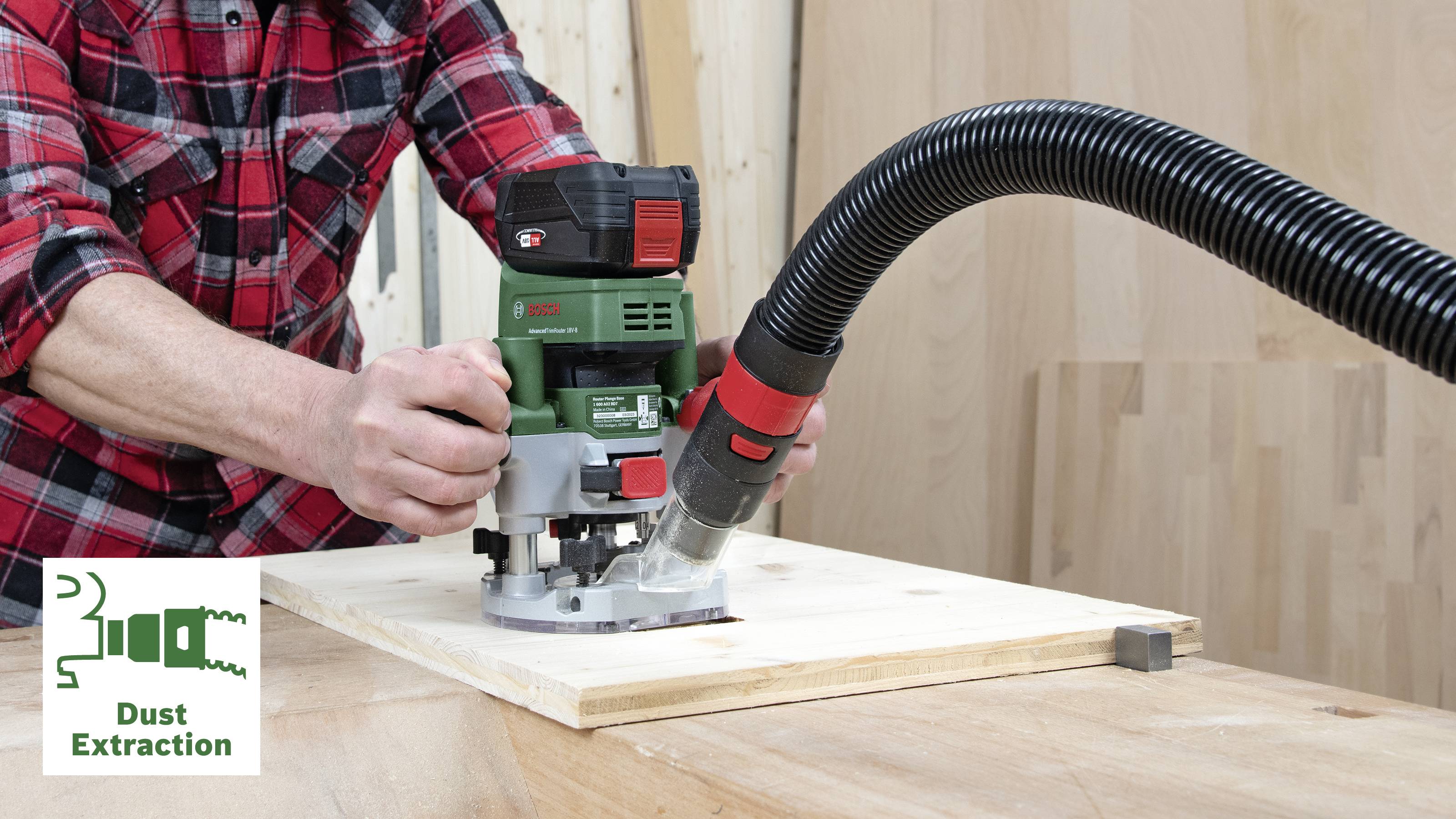 Eine Person bedient eine grüne Oberfräse auf einem Holzbrett. Ein Absaugschlauch ist an das Gerät angeschlossen.