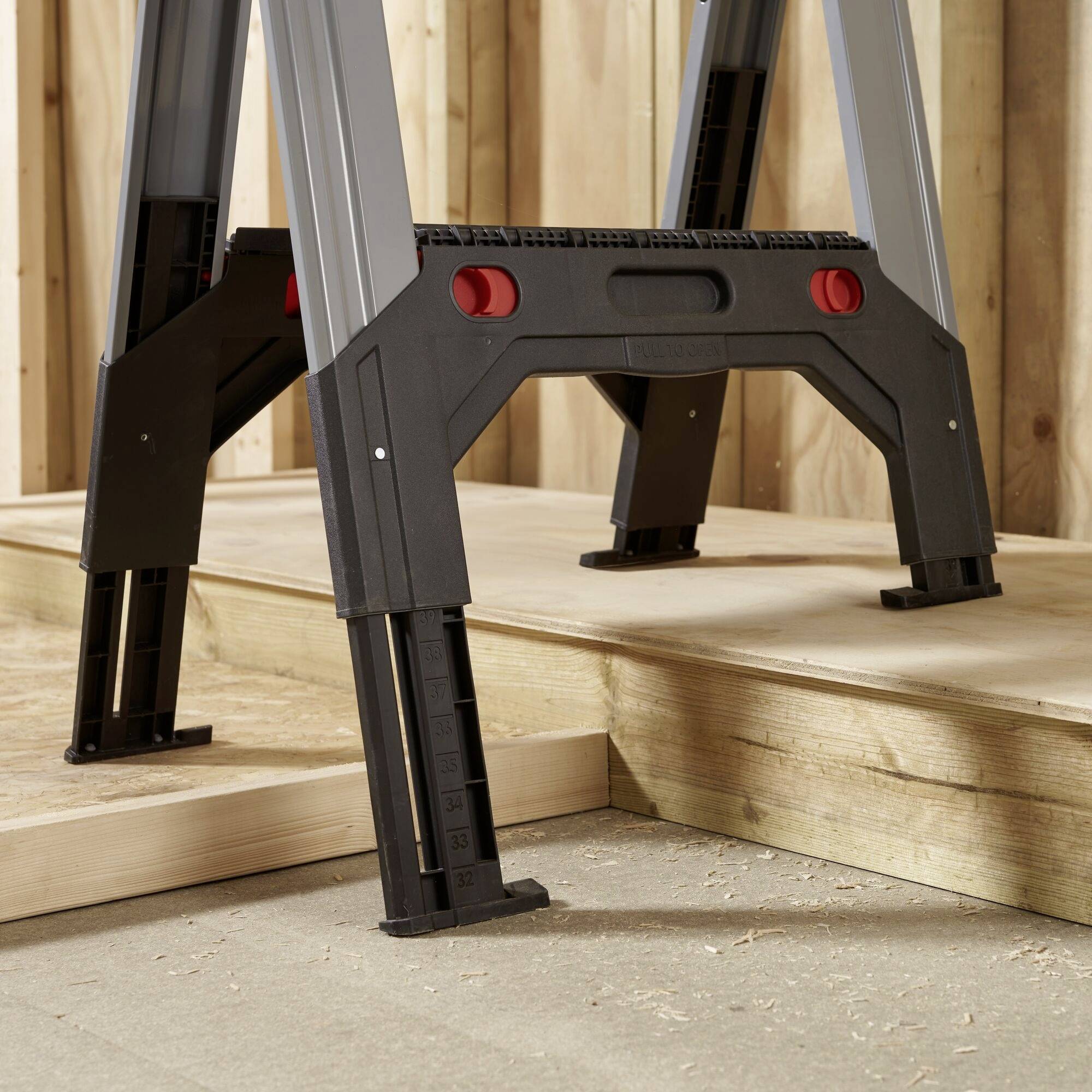 Eine faltbare Leiter aus Metall in einer Werkstattumgebung, aufgestellt auf einem Holzpodest, mit roten Verriegelungsknöpfen.