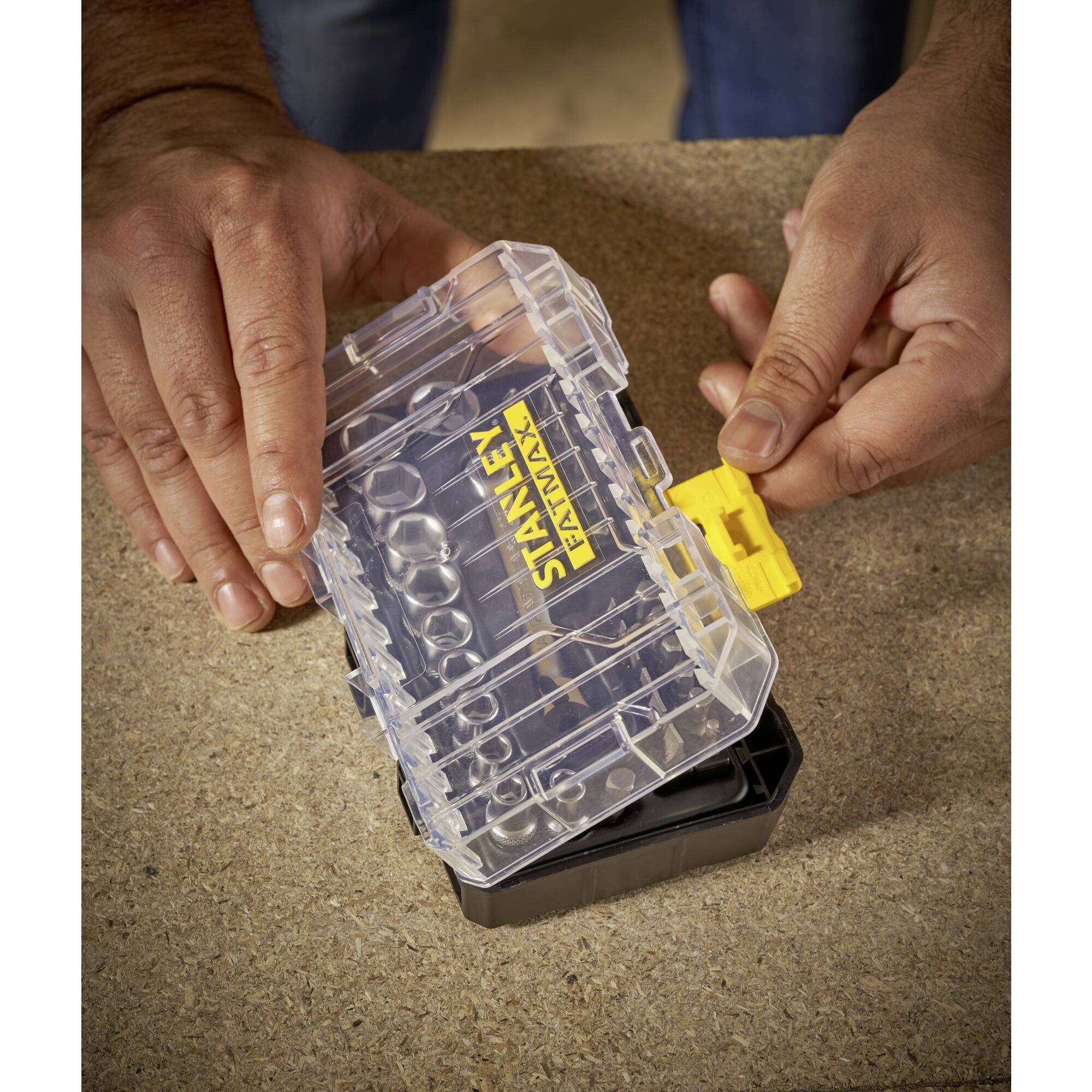 Eine Person öffnet eine kleine, transparente Kunststoffbox mit gelbem Verschluss auf einer Tischplatte.