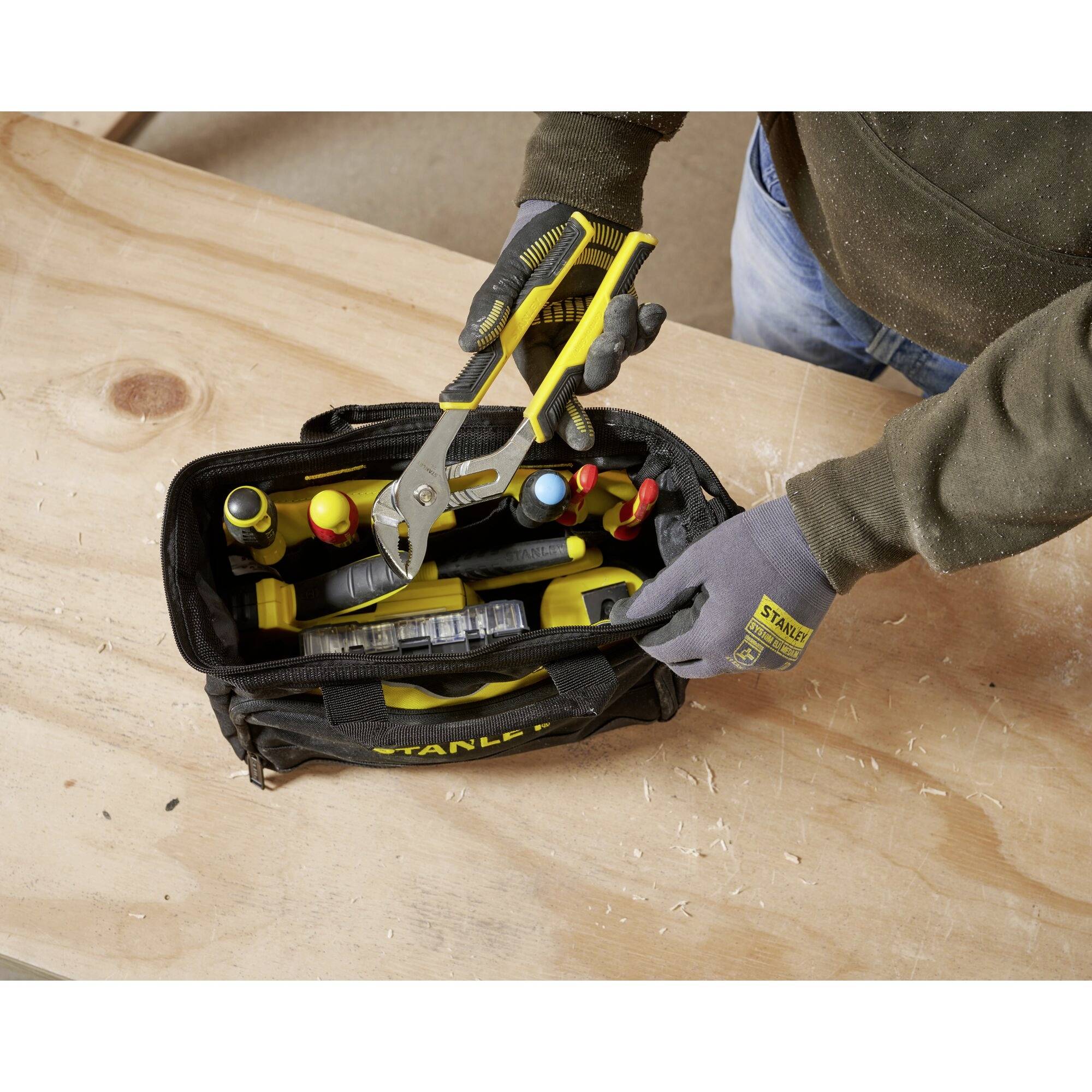 Eine Person packt eine Werkzeugtasche mit einer Zange ein. Die Tasche enthält verschiedene Werkzeuge. Sie steht auf einer Holzoberfläche.