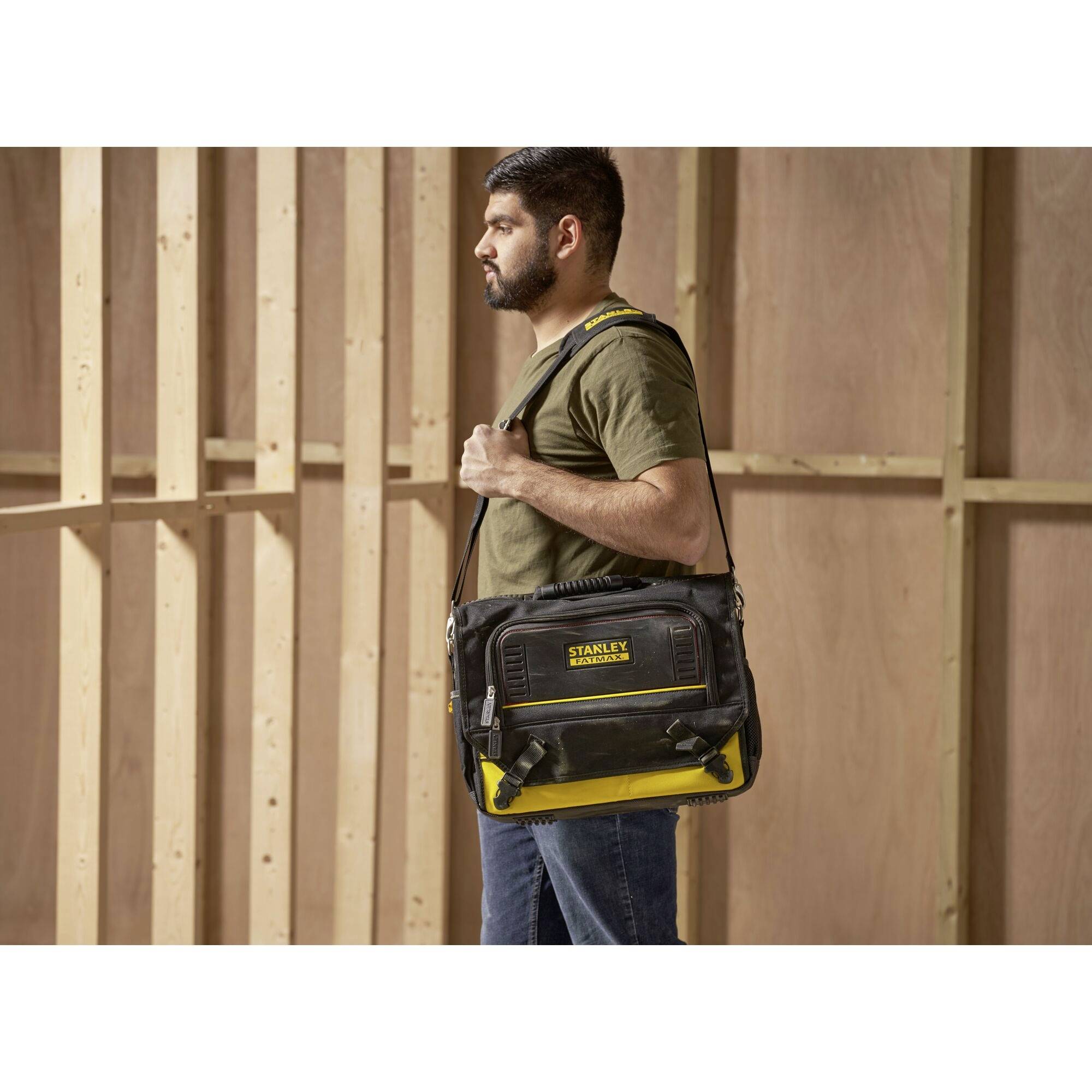 Ein Mann trägt eine schwarze und gelbe Werkzeugtasche mit der Aufschrift 'STANLEY' über der Schulter in einem unfertigen Raum.
