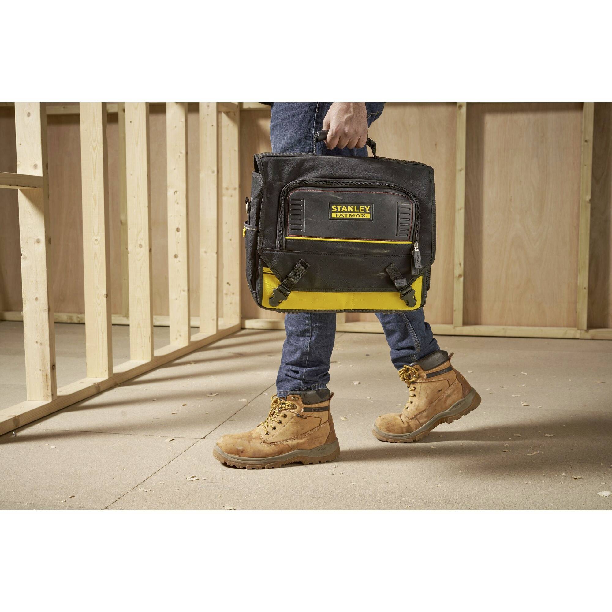 Person trägt eine schwarze, gelbe Werkzeugtasche in einem unfertigen Holzrahmenbau. Die Tasche ist mit 'STANLEY FATMAX' beschriftet.