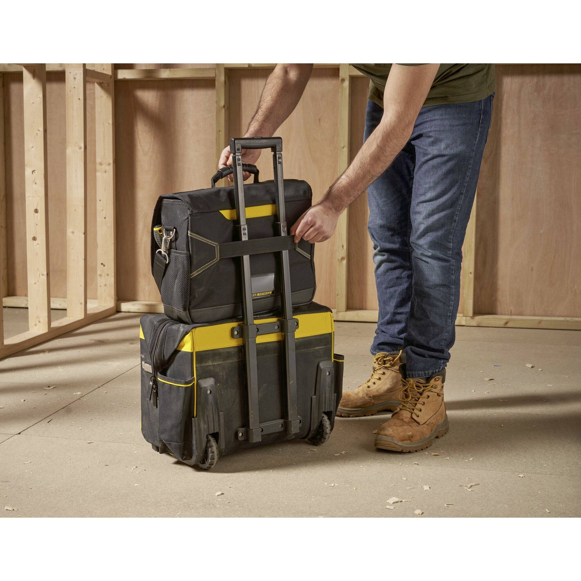 Eine Person in Baukleidung transportiert zwei gestapelte Werkzeugtaschen auf Rollen in einem unfertigen Holzrahmengebäude.