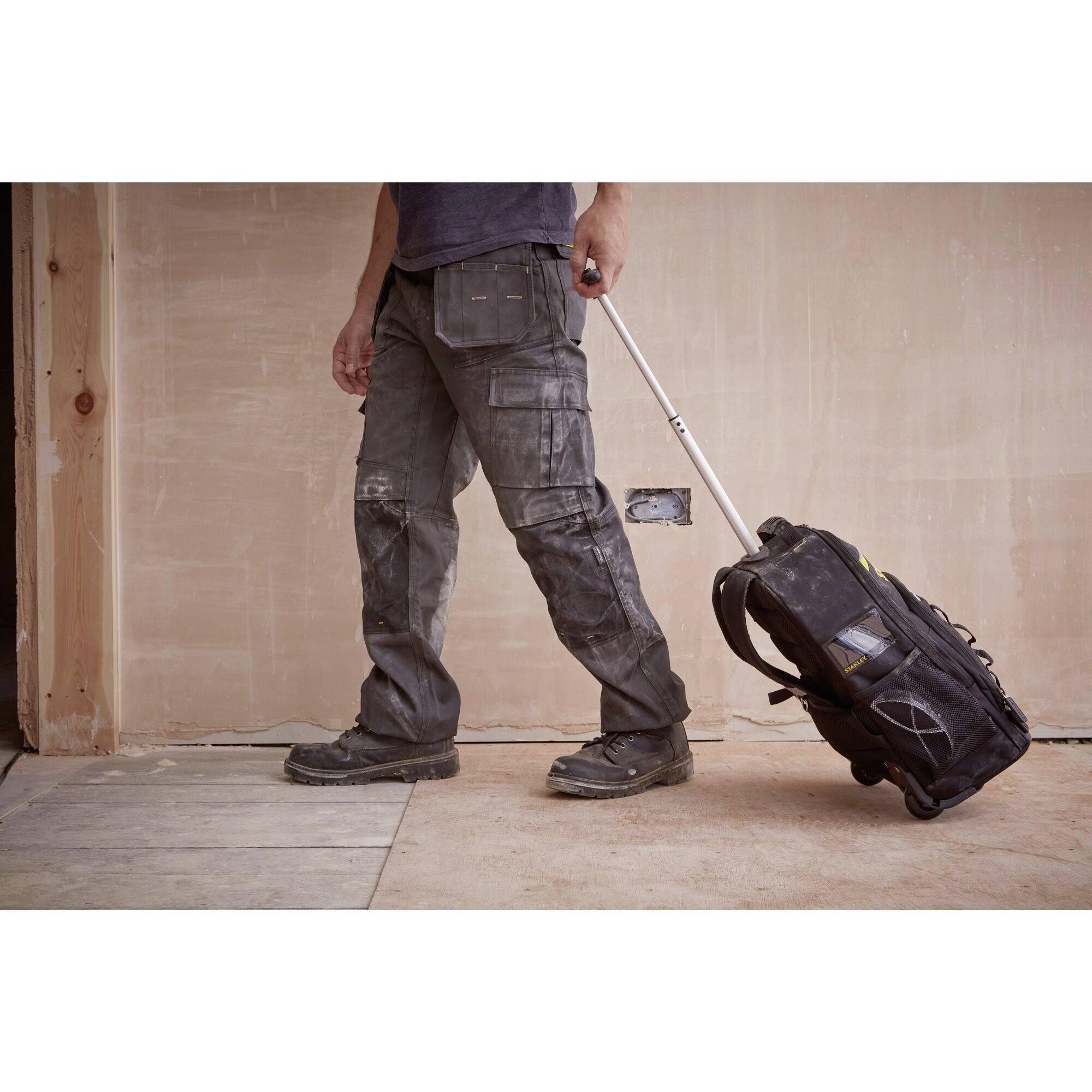 Eine Person zieht einen Rucksack mit Rädern über einen Bauholzboden. Die Person trägt Arbeitskleidung und ist von der Taille abwärts sichtbar.