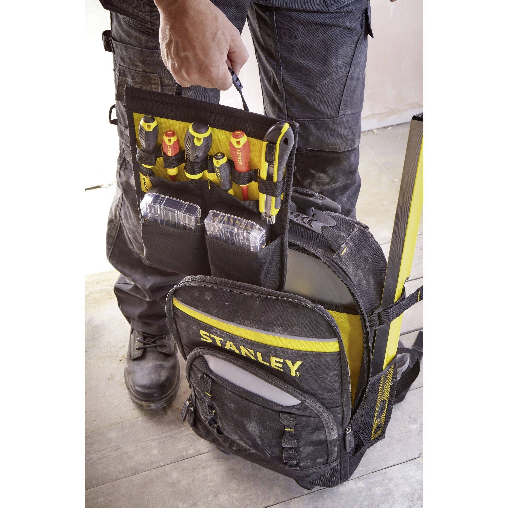 Eine Person hält einen Werkzeugrucksack mit mehreren Werkzeugfächern. Schraubendreher und kleine Boxen sind sichtbar.