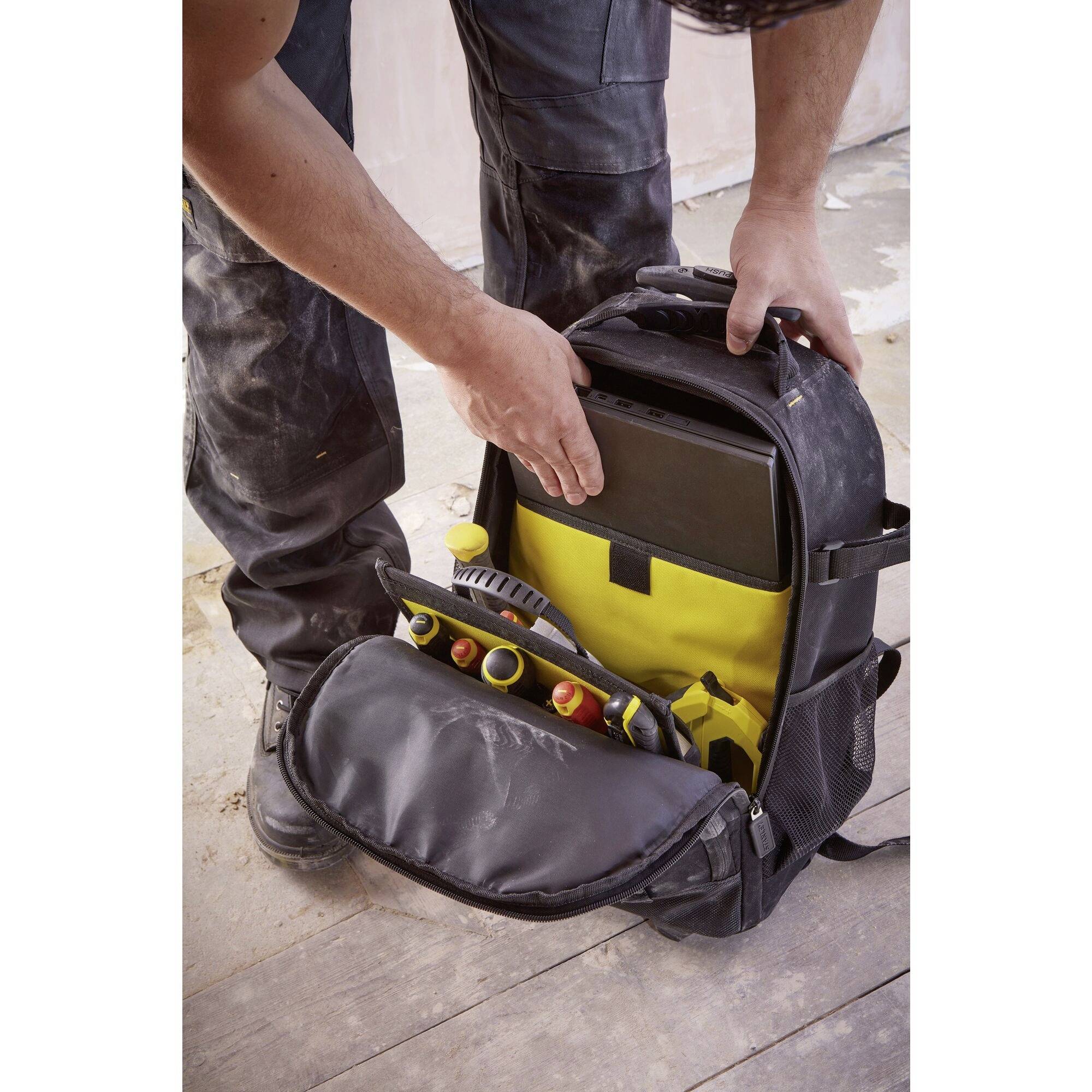 Eine Person in Arbeitskleidung entnimmt ein Tablet aus einem teilweise geöffneten Werkzeugrucksack. Im Rucksack befinden sich auch Werkzeuge.