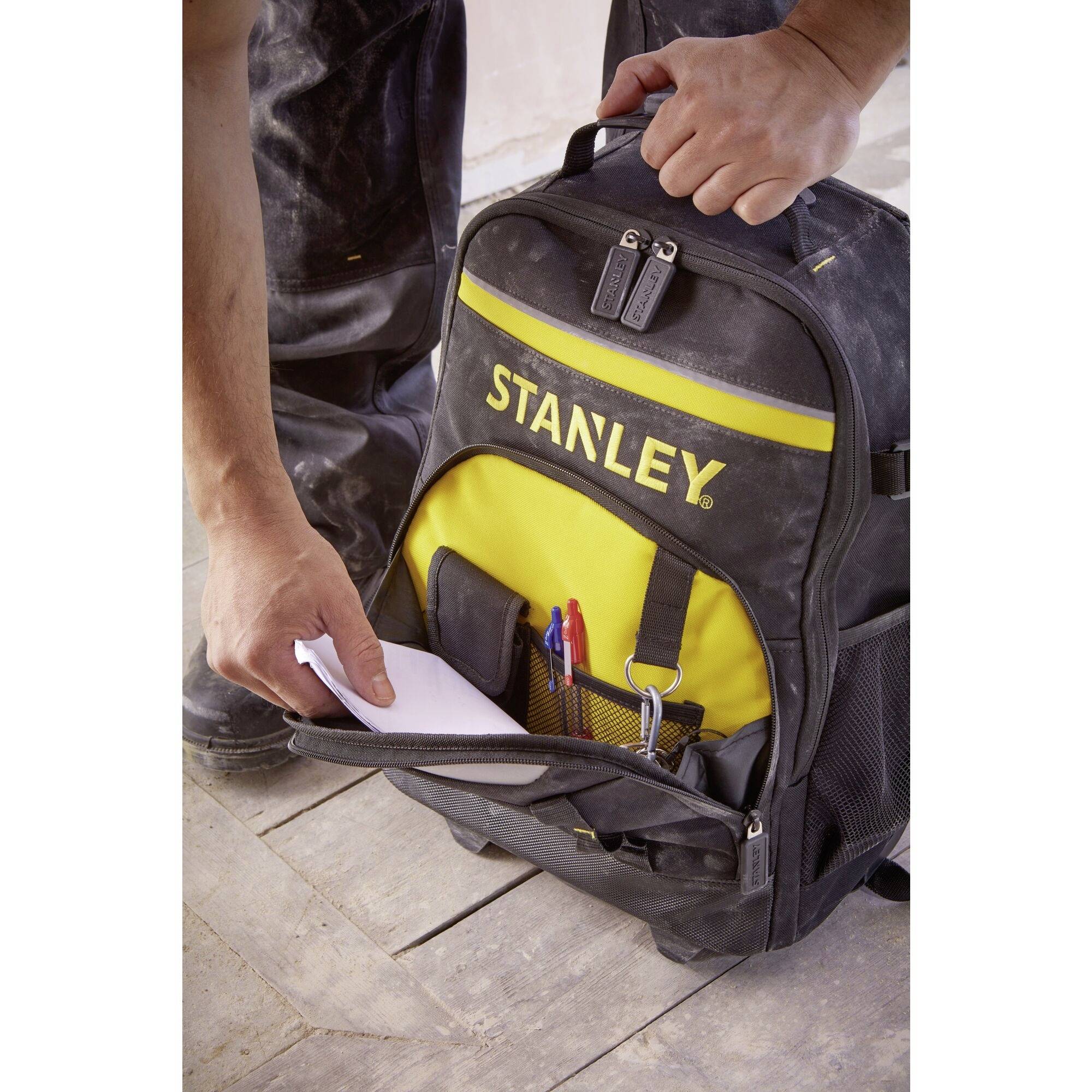 Eine Person legt ein Papier in einen schwarz-gelben Stanley-Werkzeugrucksack, der auf einem Holzboden steht.