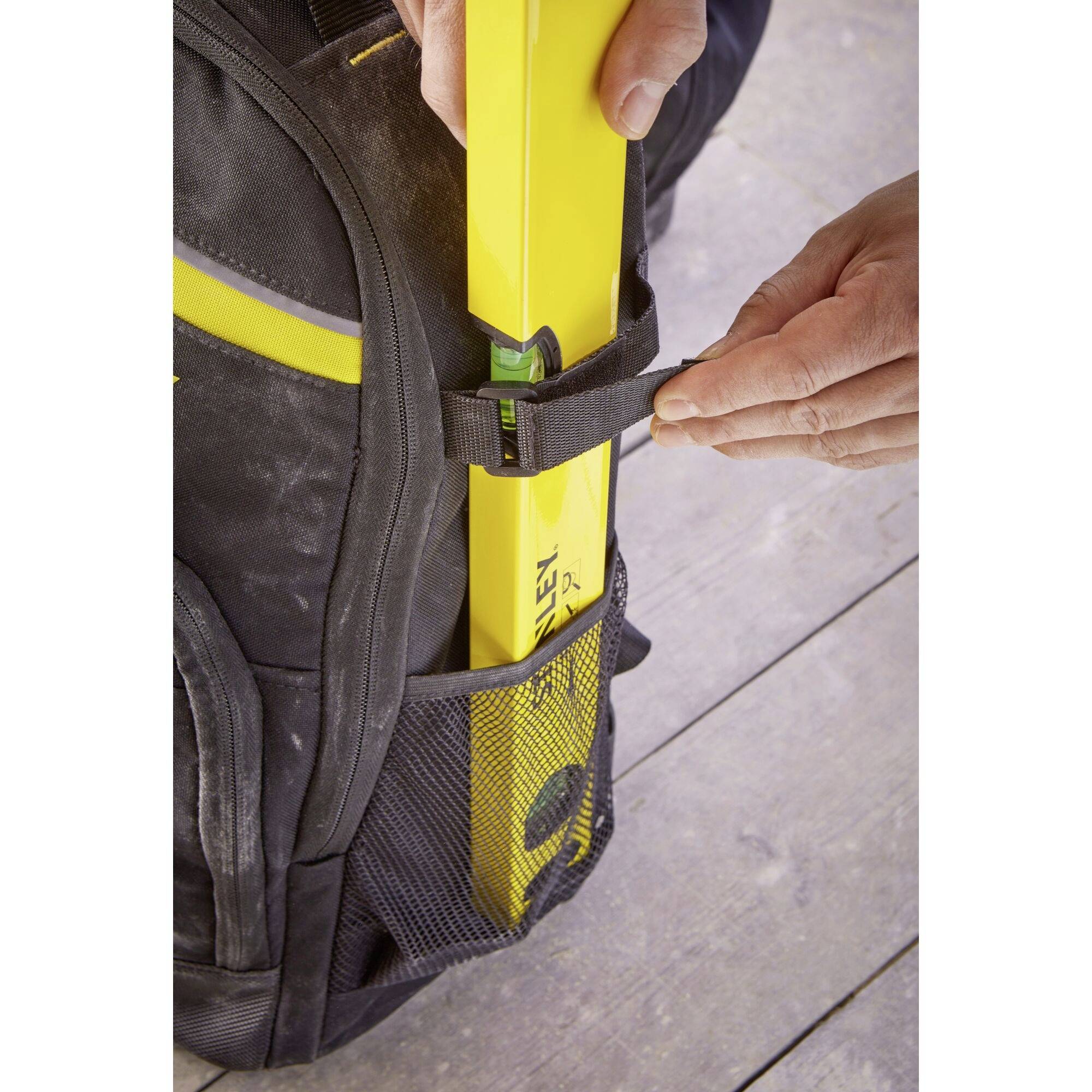 Hand befestigt Wasserwaage an einem schwarzen Rucksack; die Wasserwaage wird in einer seitlichen Netztasche des Rucksacks verstaut.
