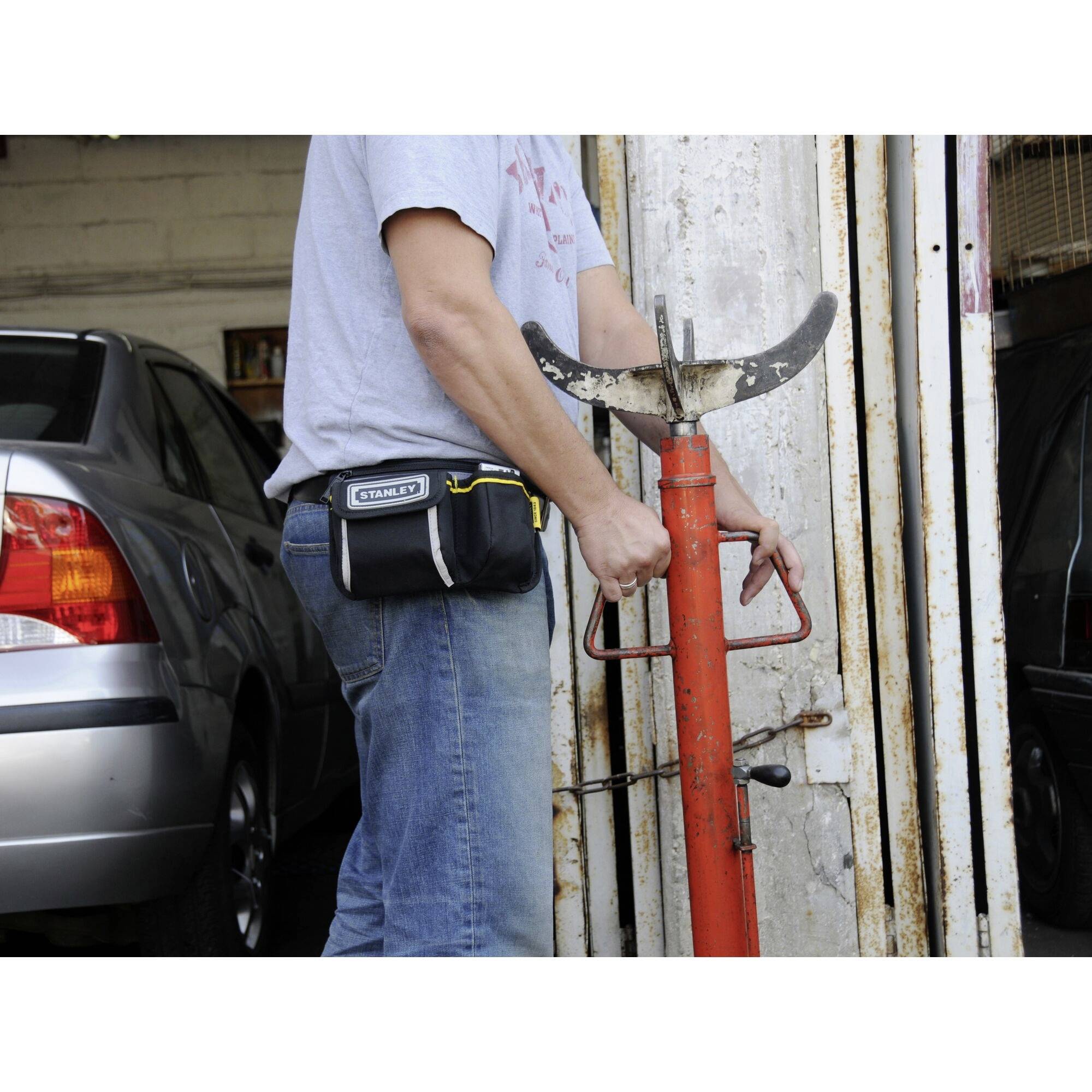 Eine Person in Jeans und T-Shirt nutzt ein rotes Werkzeug, vermutlich ein Wagenheber, in einer Autowerkstatt neben einem geparkten Auto.