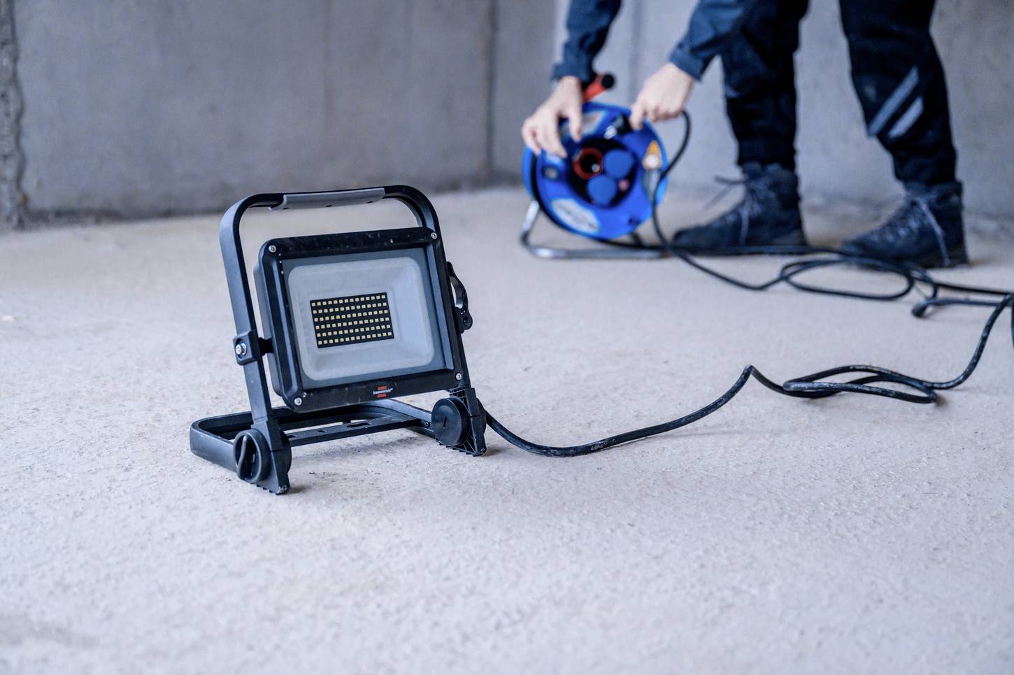 Ein Baustrahler auf dem Boden ist über ein langes Kabel mit einer Kabeltrommel verbunden. Eine Person im Hintergrund hält die Trommel.