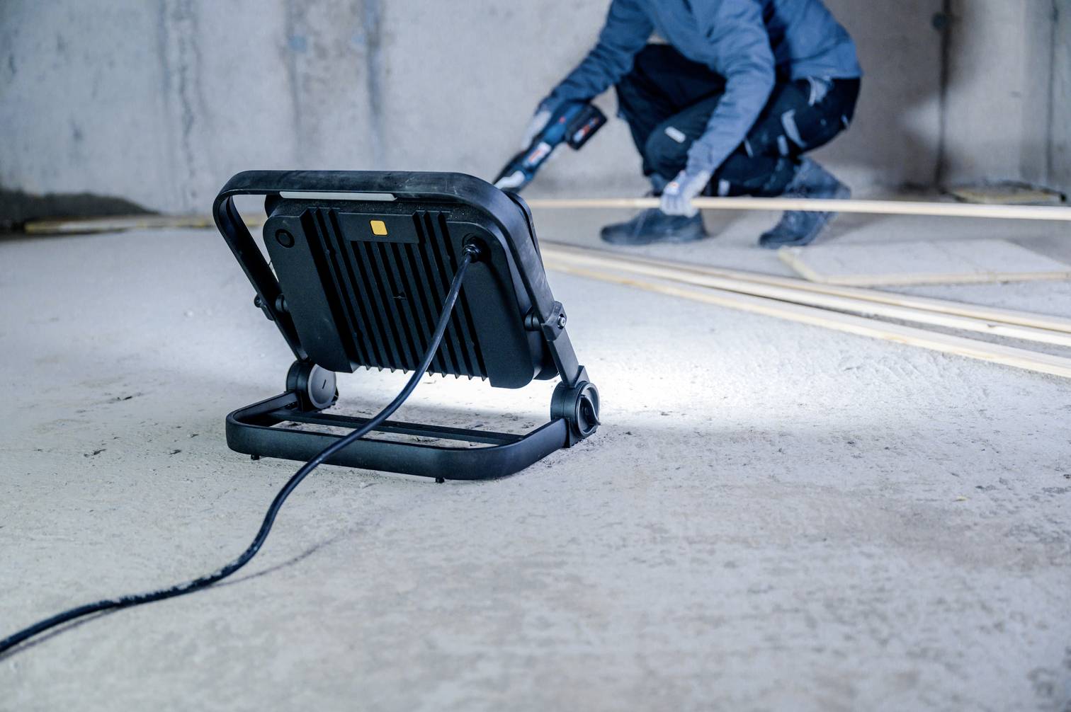Eine Person in Arbeitskleidung nutzt ein Werkzeug in einem unfertigen Raum. Im Vordergrund steht ein Arbeitslicht, das den Bereich beleuchtet.
