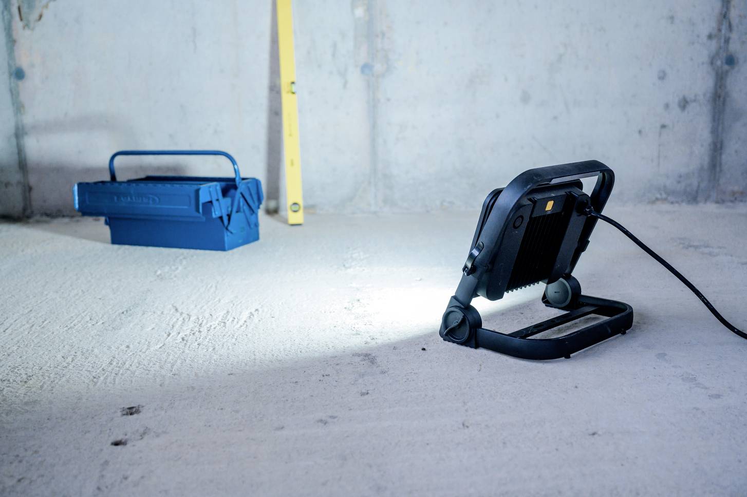 Blaue Werkzeugkiste und Arbeitsleuchte auf einem Betonboden in einem unfertigen Raum. Die Leuchte wirft Licht auf den Boden.