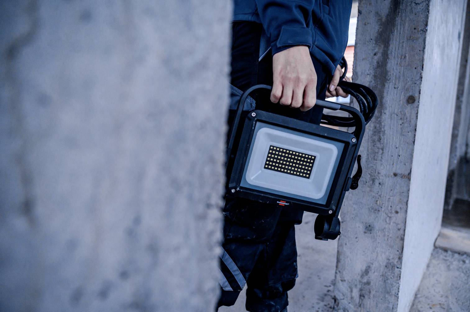 Eine Person hält einen tragbaren LED-Baustrahler. Der Hintergrund zeigt Betonwände, was auf eine Baustelle hindeutet.
