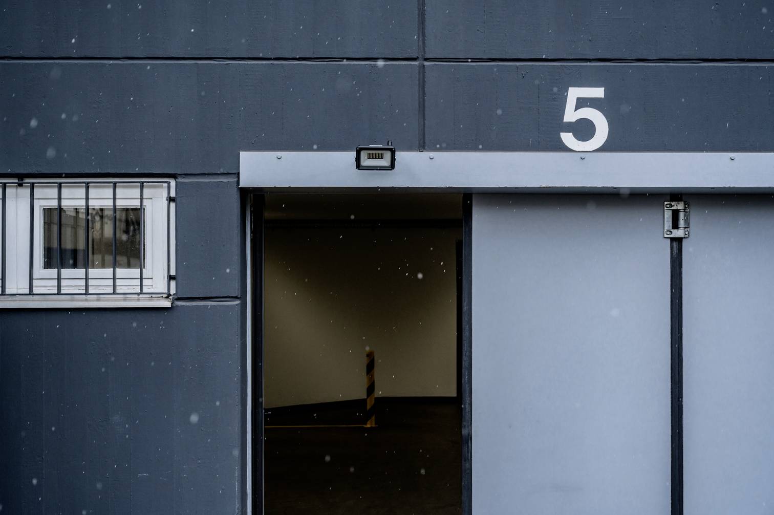 Ungeöffnete graue Schiebetür mit der Zahl 5, daneben kleines Fenster mit Gitter. Trotz des Schneefalls bietet das Bild eine ruhige Atmosphäre.