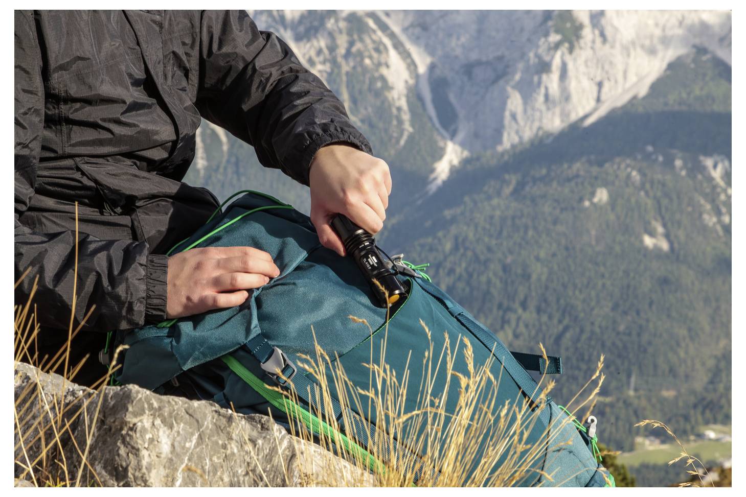 Person, die eine tragbare Taschenlampe auf einen blaugrünen Rucksack richtet, während sie auf Steinen in den Bergen sitzt, mit alpinen Gipfeln im Hintergrund.