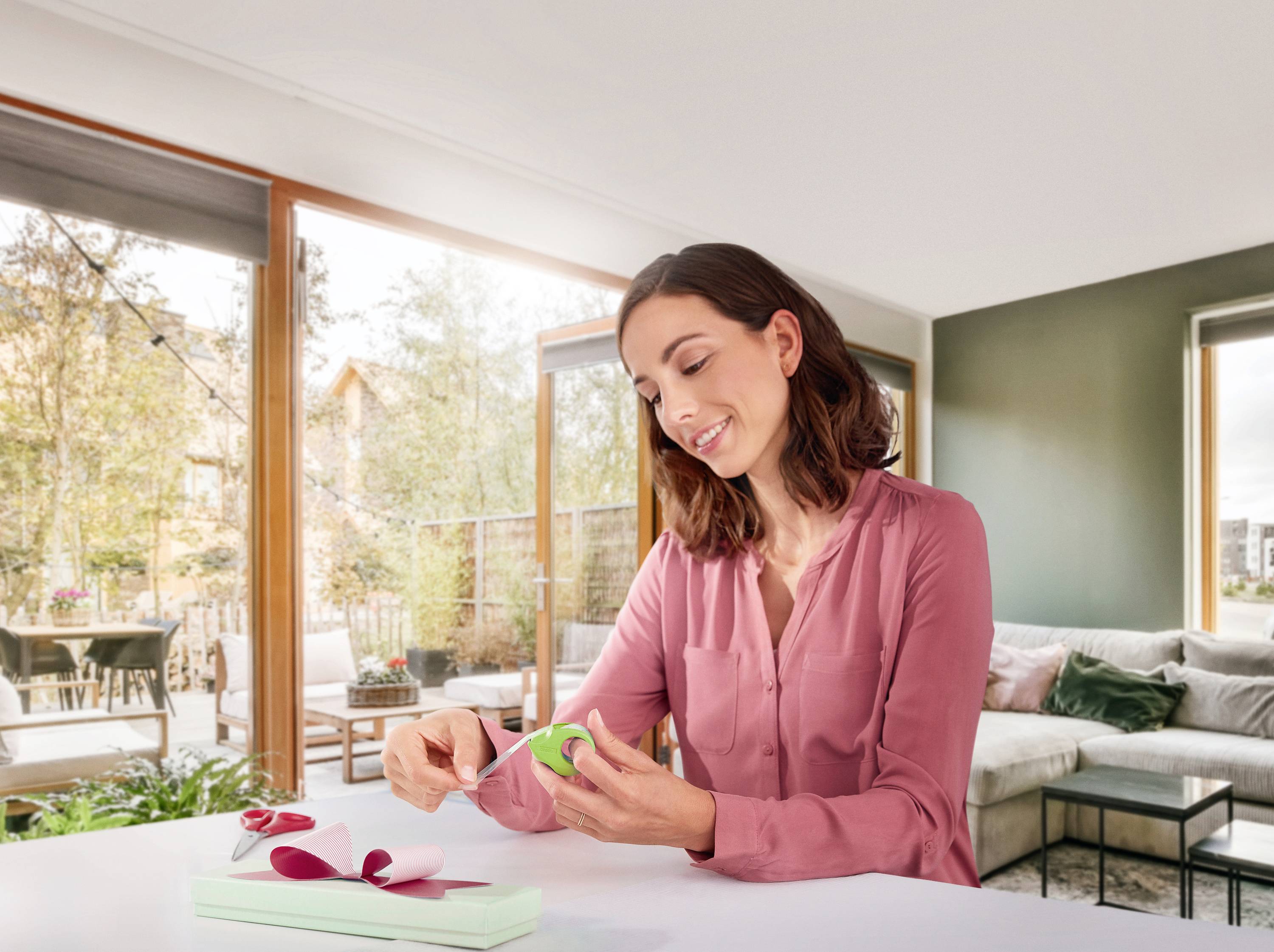 Eine Person in einem modernen Wohnzimmer öffnet ein Geschenk in einer grünen Box mit rosa Band. Im Hintergrund große Fenster und Pflanzen.