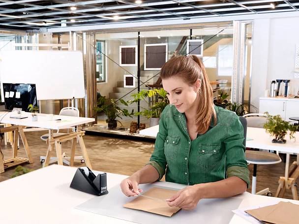 Eine Frau in einem grünen Hemd sitzt an einem Schreibtisch in einem modernen Büro und steckt einen Brief in einen Umschlag. Sie ist konzentriert bei ihrer Aufgabe.