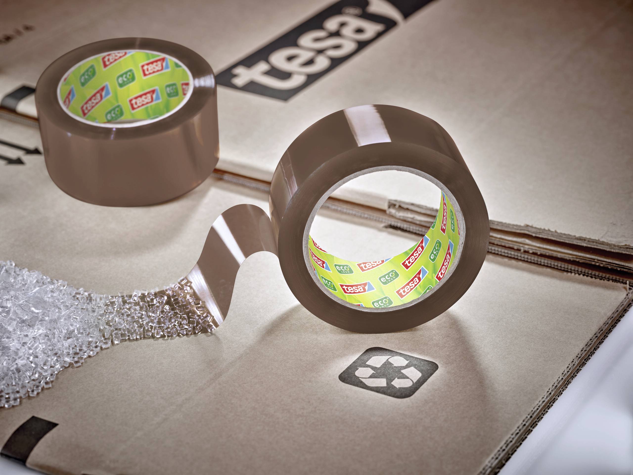 Zwei Rollen braunes Verpackungsklebeband, eine mit abgezogenem Band, liegen auf Karton. Im Hintergrund ist das Logo 'tesa' sichtbar.