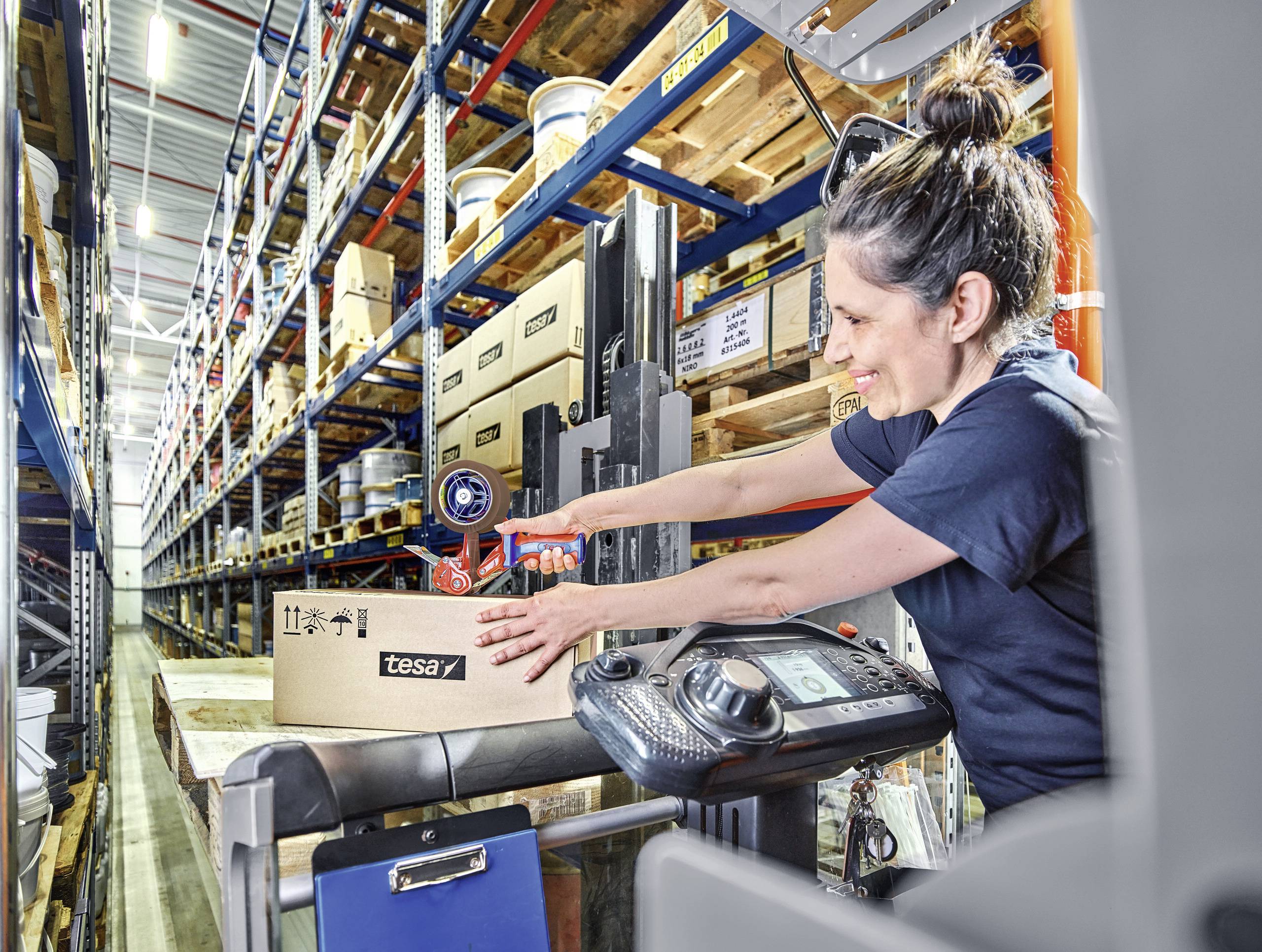 Eine Frau bedient einen Gabelstapler in einem Lager. Sie stapelt Kartons, während sie das Fahrzeug steuert. Regale voller Kartons im Hintergrund.