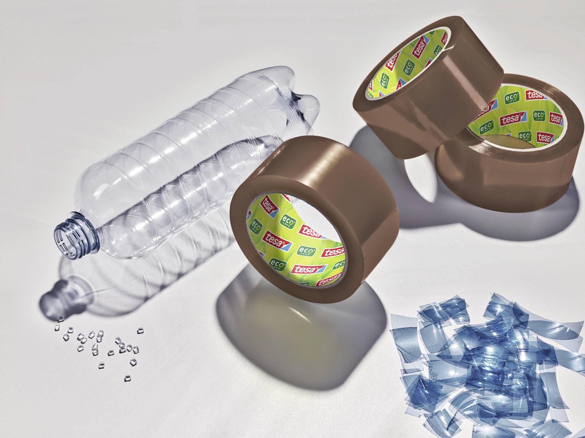 Leere Plastikflasche, drei Rollen Klebeband und Kunststoffstücke auf einem Tisch. Darstellung von Recycling oder Kunststoffverarbeitung.