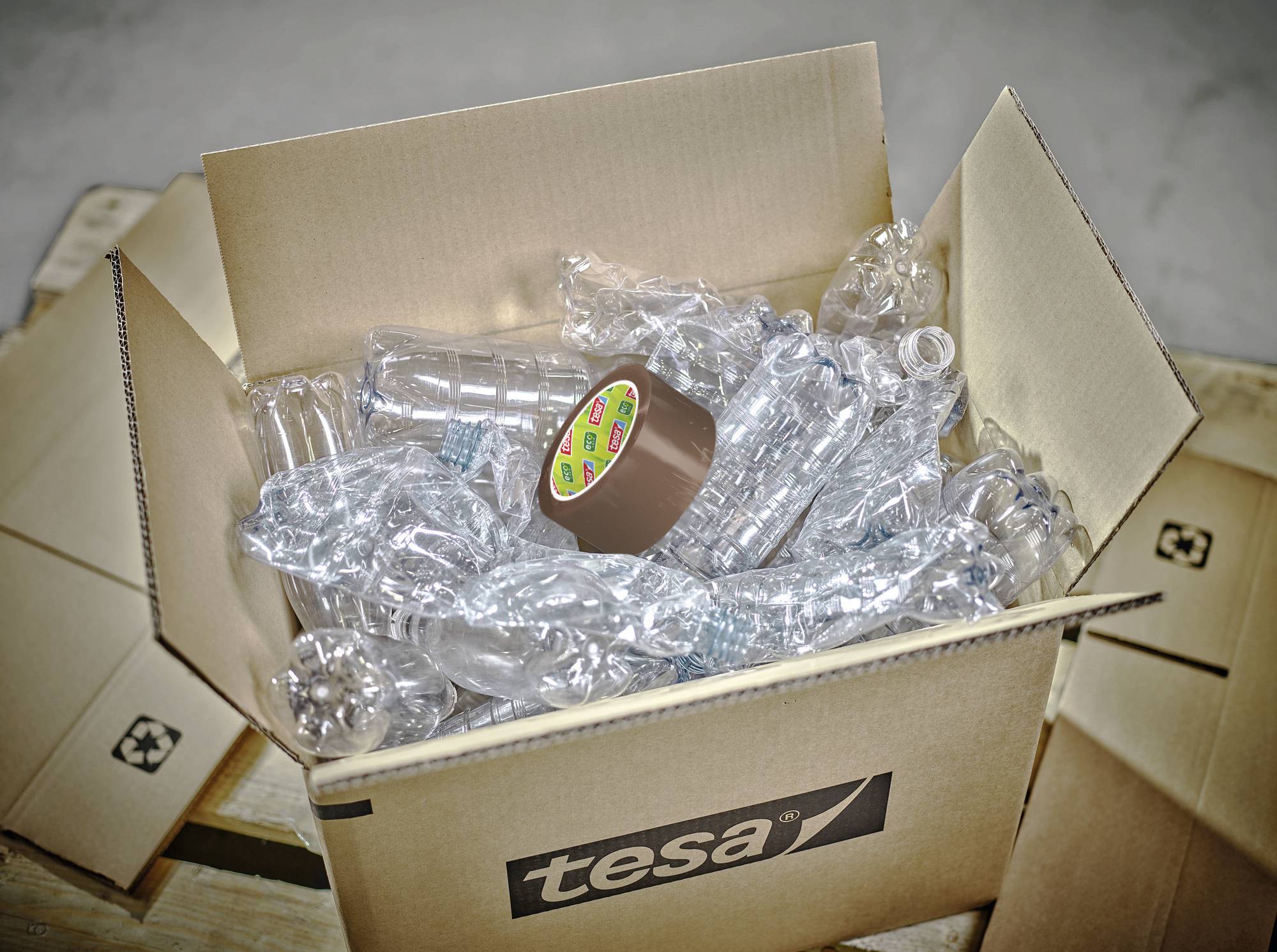 Ein offener Karton mit zerdrückten Plastikflaschen und einer Rolle braunem Klebeband, auf einem Holzpalettenset.