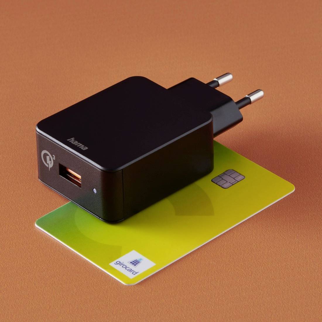 Ein schwarzes USB-Ladegerät liegt auf einer gelben Girocard mit Chip, auf einem orangefarbenen Untergrund.