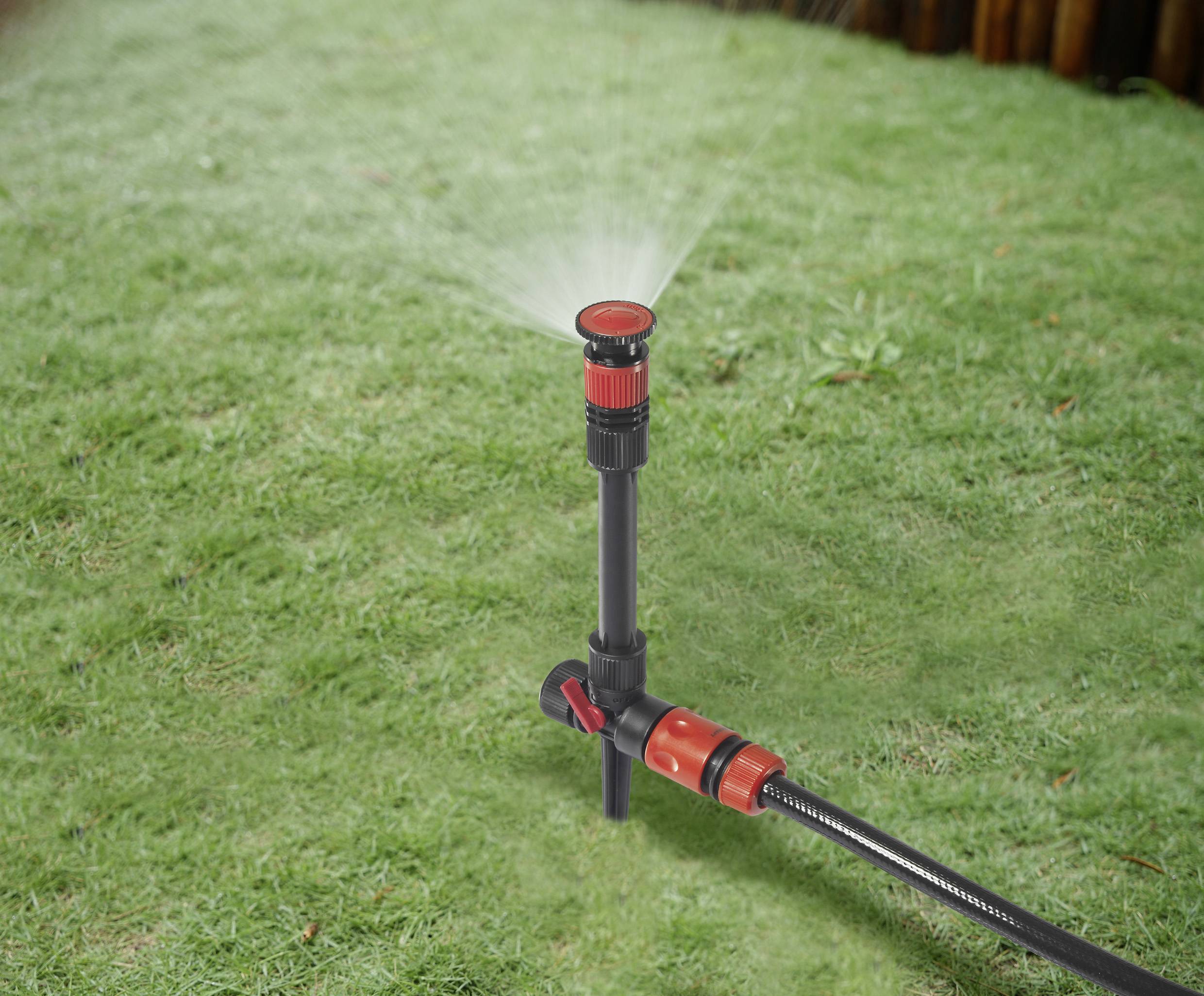 Ein Rasensprenger auf einer Rasenfläche verteilt Wasser gleichmäßig. Der Sprühkopf ist rot und schwarz, verbunden mit einem Wasserschlauch.