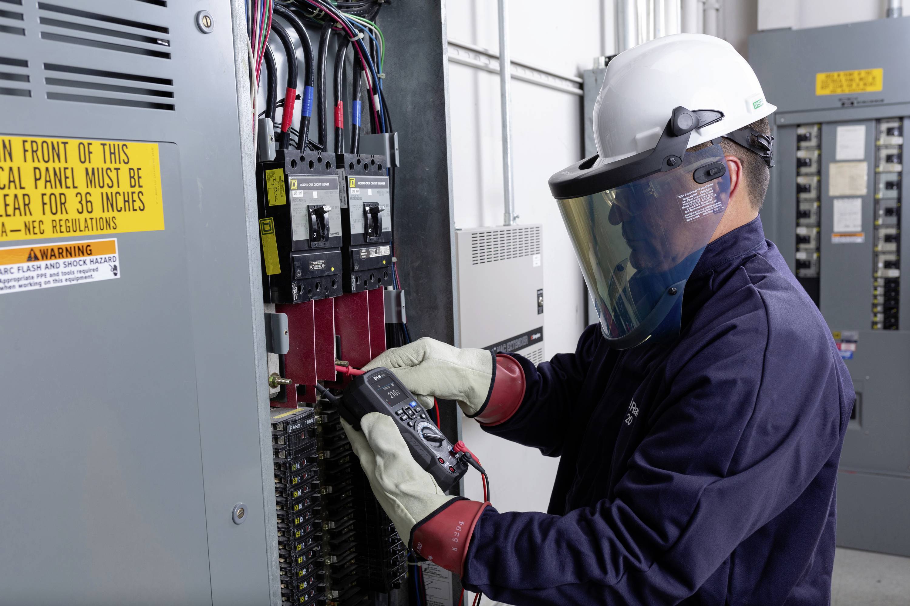 Ein Techniker in Schutzkleidung misst mit einem Gerät in einem elektrischen Schaltschrank. Sicherheitswarnungen sind sichtbar.