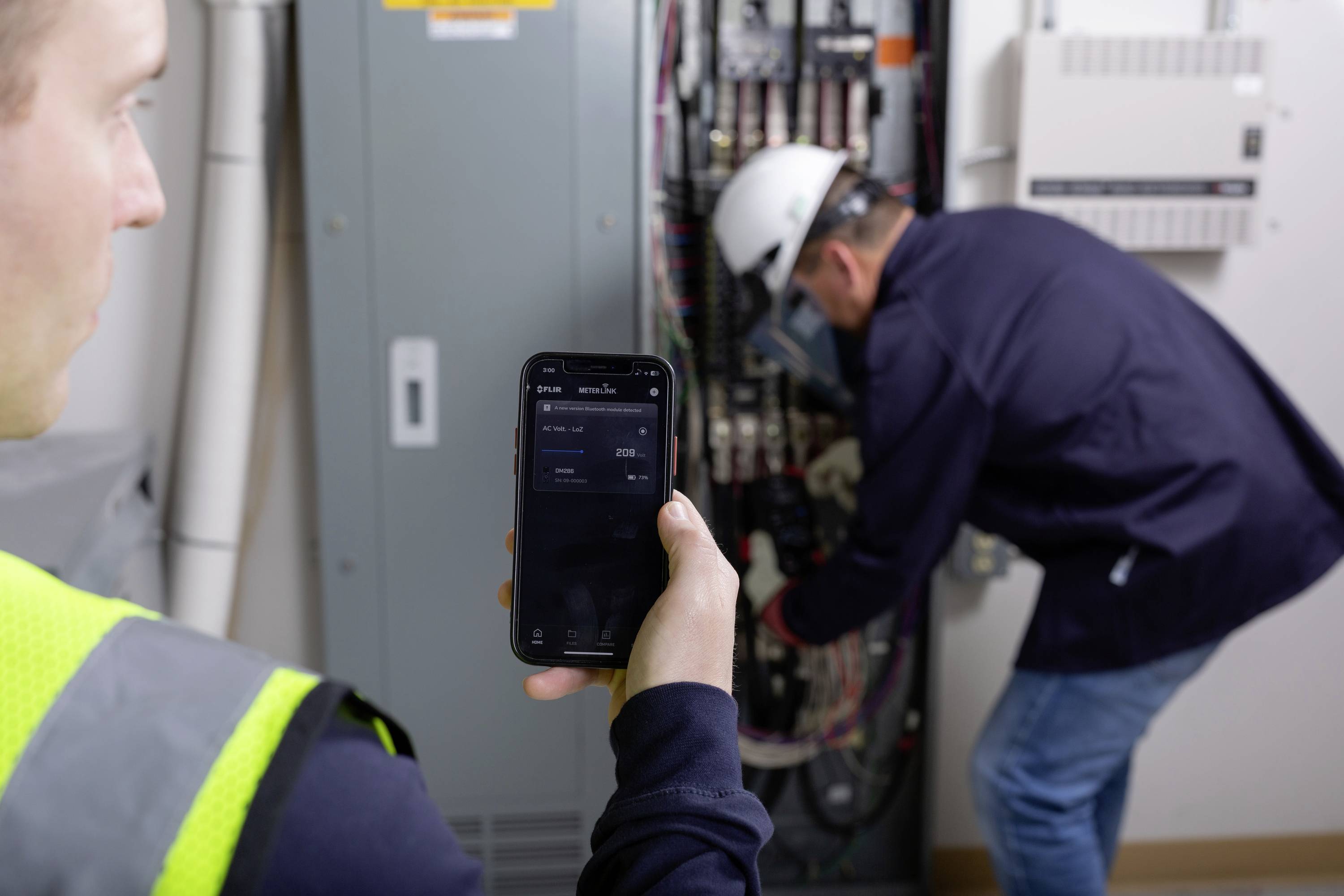 Ein Arbeiter steht vor einem geöffneten Schaltkasten und benutzt ein Smartphone, um den Stromverbrauch zu prüfen. Ein Techniker arbeitet im Hintergrund.