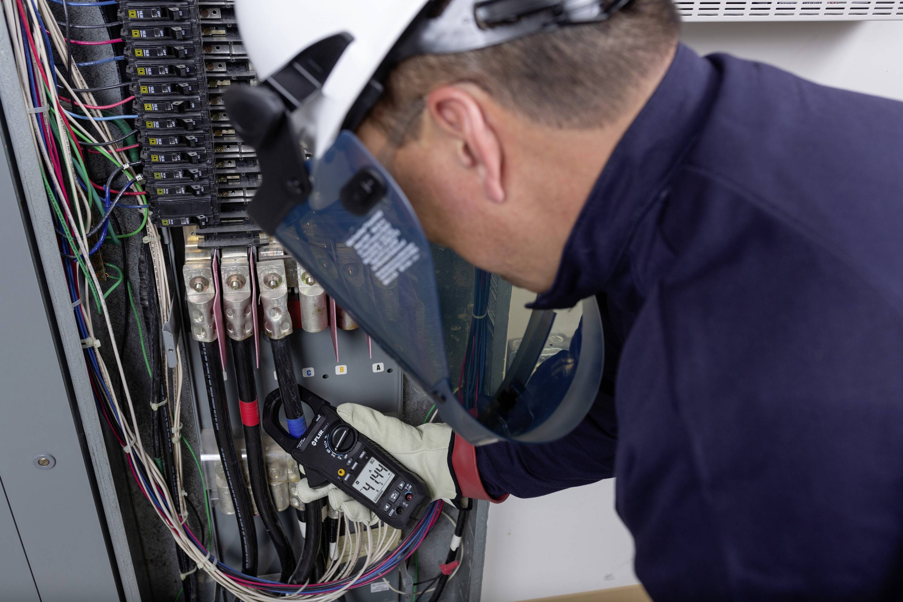 Ein Elektriker in Schutzkleidung misst Spannung in einem Schaltschrank mit einem Multimeter. Sichtbar sind viele bunte Kabel.