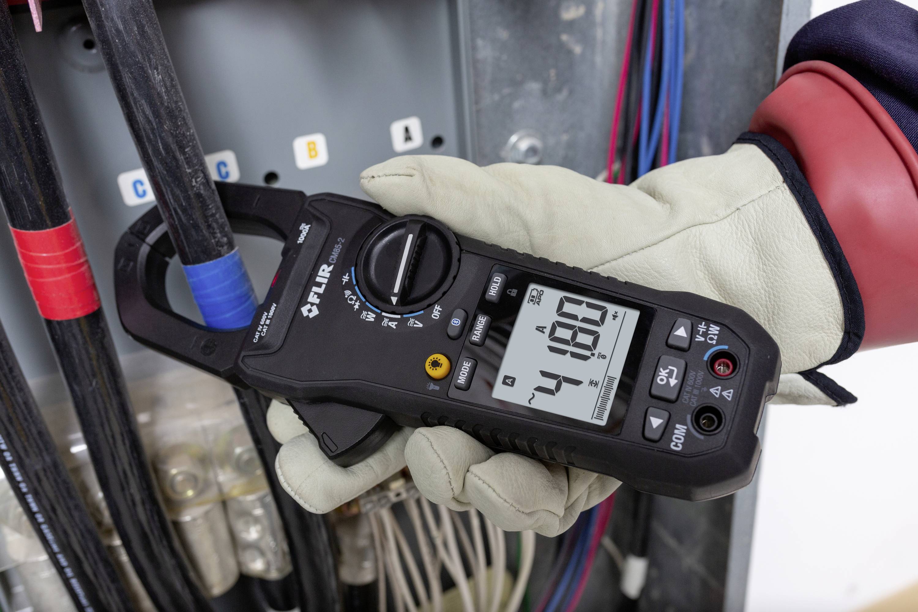 Eine behandschuhte Hand hält ein digitales Multimeter, das 48,0 Ampere misst, vor einem elektrischen Verteilerkasten.