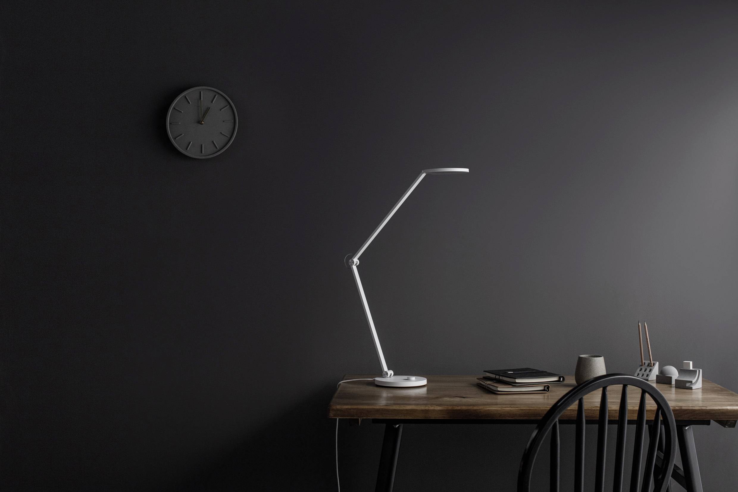 Arbeitsplatz mit dunklem Holztisch, minimalistischer weißer Tischlampe und Wanduhr, in einem schlicht eingerichteten Raum.