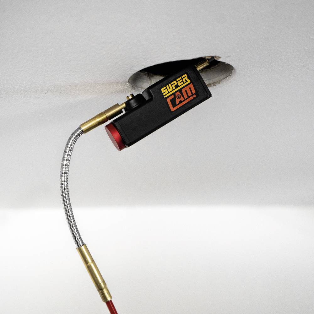 Ein flexibles Inspektionswerkzeug mit Kamera schaut durch ein Loch in der Wand. Auf der Kamera steht 'SUPER CAM'.