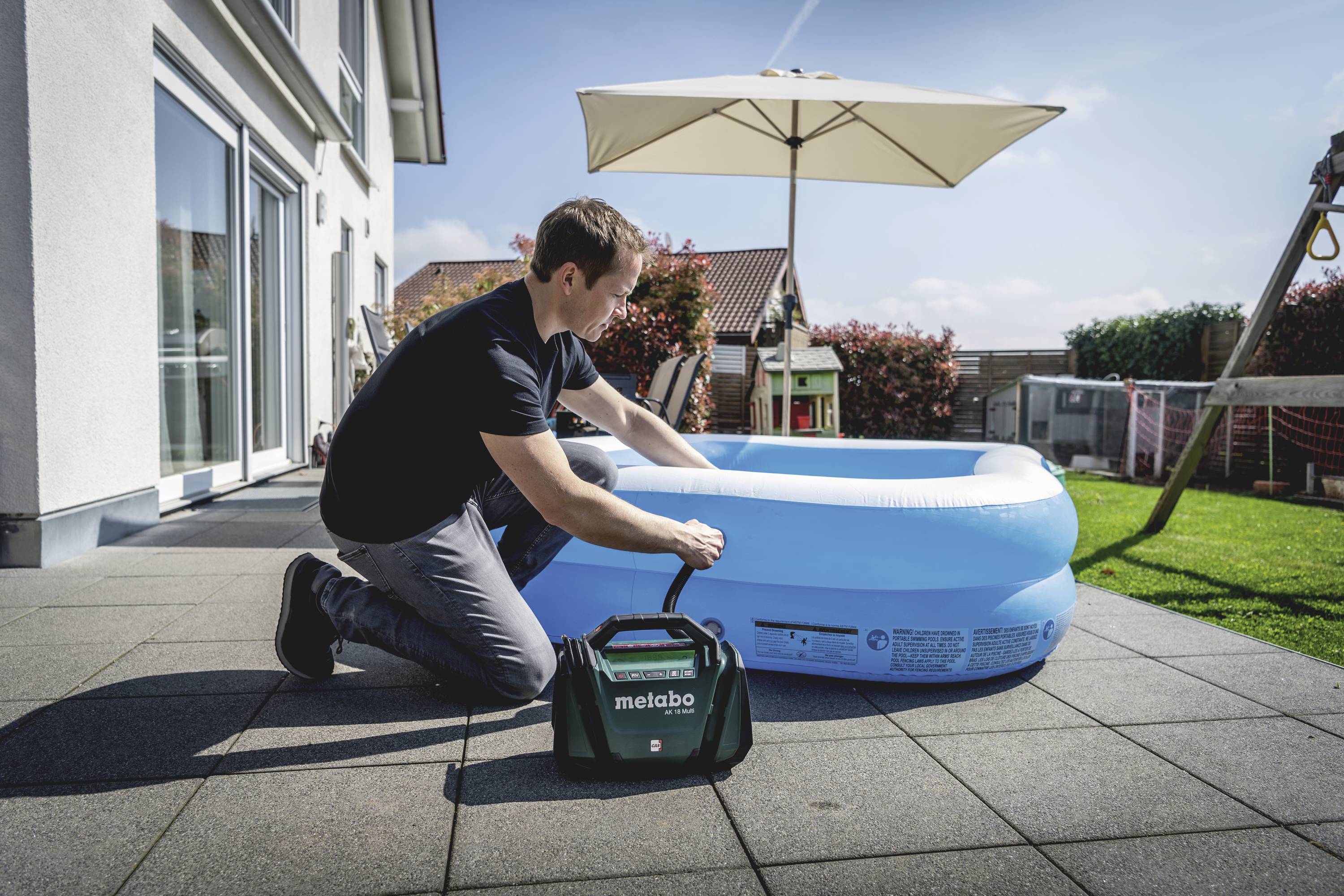 Ein Mann pumpt auf einer Terrasse ein aufblasbares rundes Kinderplanschbecken mit einer elektrischen Pumpe auf. Im Hintergrund ist ein Garten mit Sonnenschirm zu sehen.