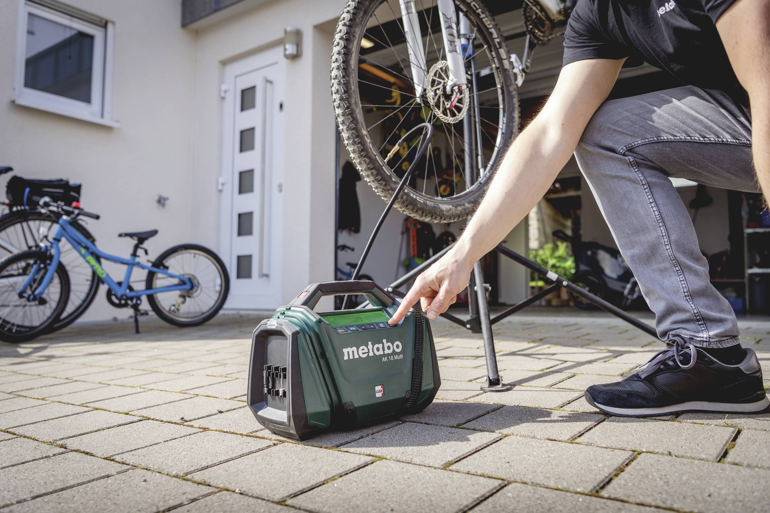 Ein Mann kniet neben einem tragbaren Aktenkoffer mit der Aufschrift 'metabo'. Im Hintergrund sind zwei Fahrräder und eine Garage zu sehen.