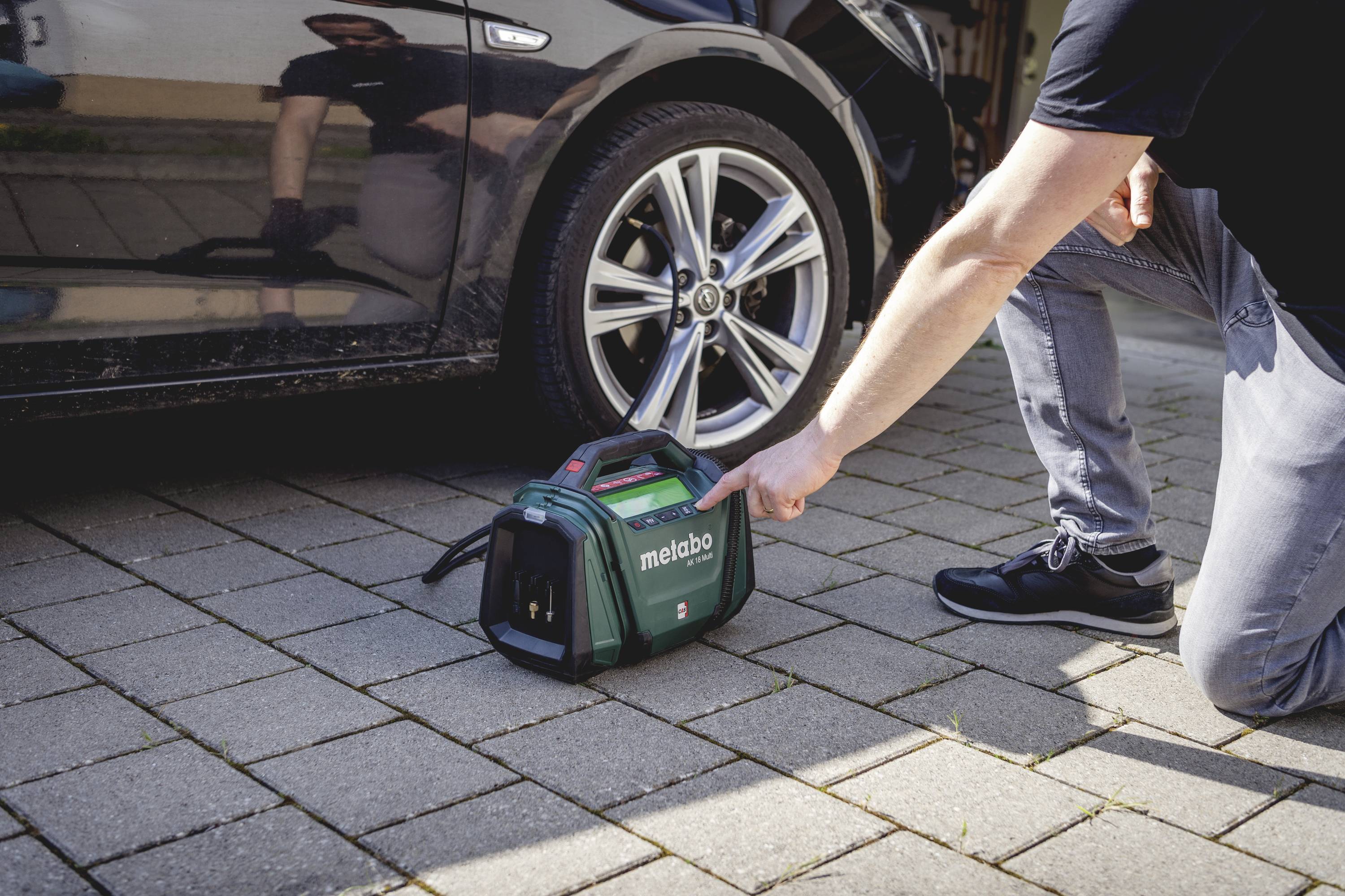 Ein Mann zeigt auf einen Werkzeugkoffer der Marke 'Metabo' auf dem Boden neben einem geparkten Auto.