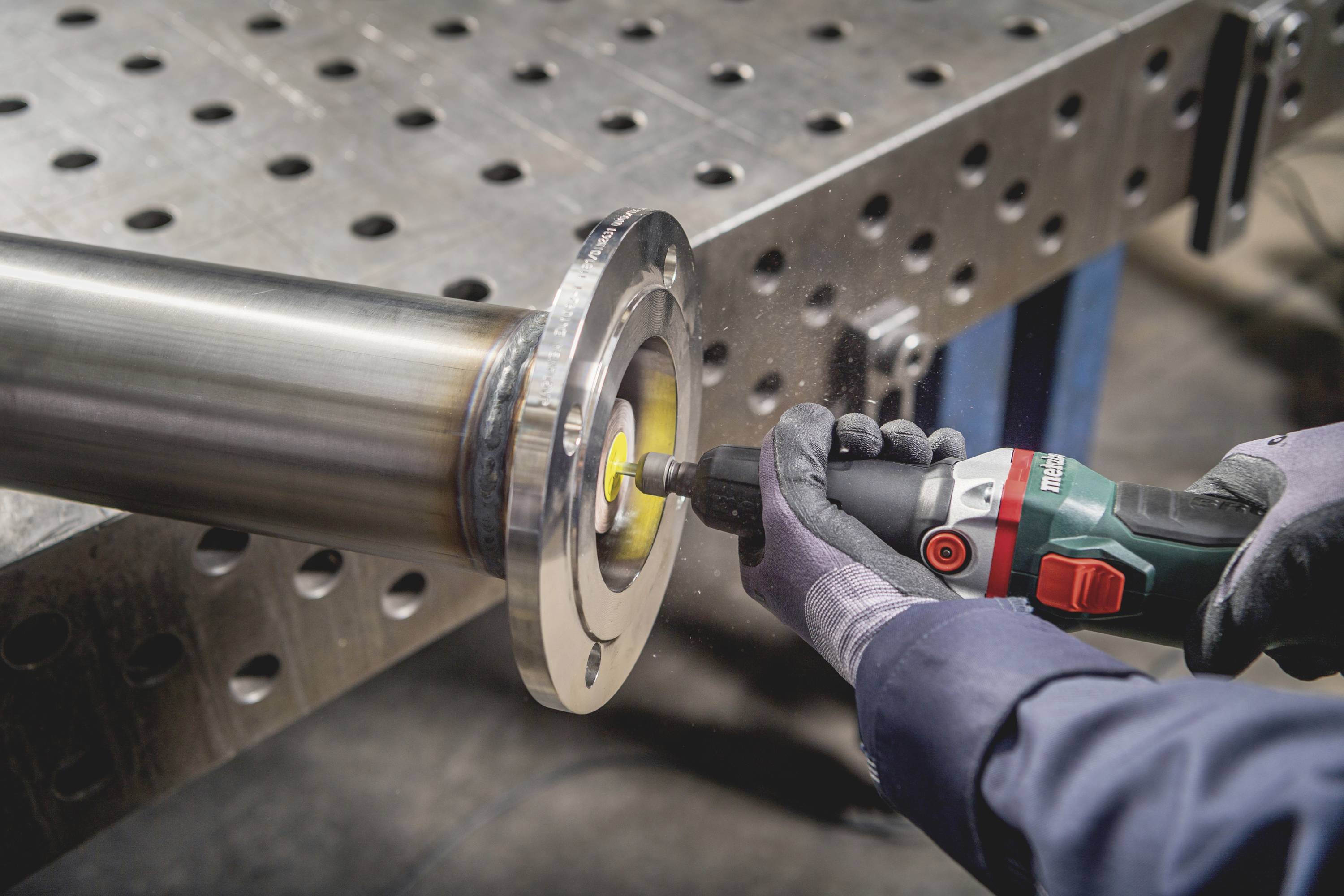 Eine Person schleift den Rand eines Metallrohrs mit einem elektrisch betriebenen Winkelschleifer auf einem Arbeitstisch.