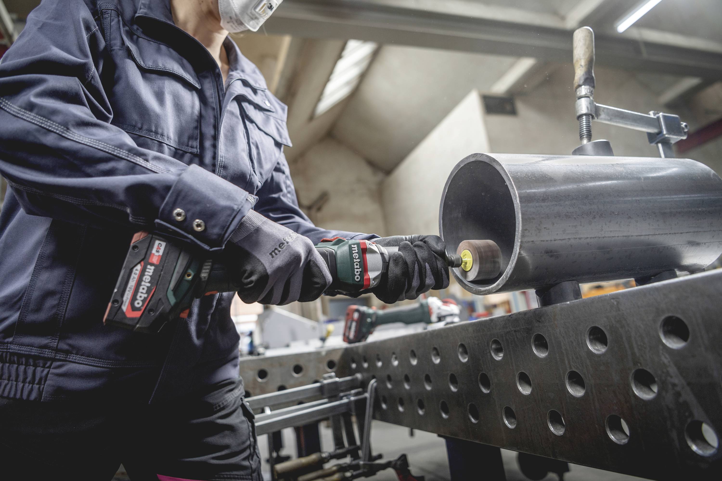 Eine Person in Arbeitskleidung schleift ein Metallrohr mit einem Handwerkzeug auf einem Werkstatttisch.