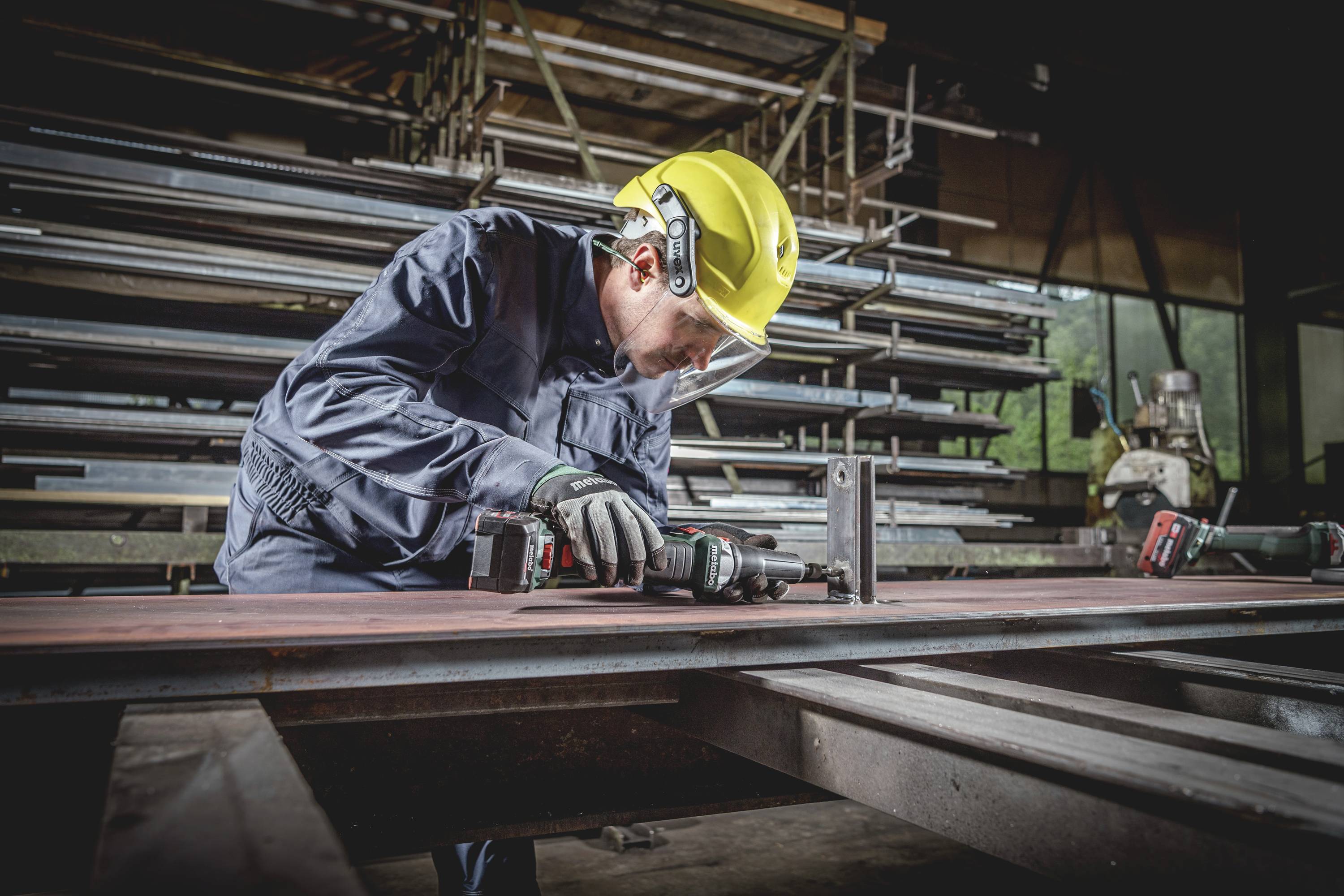 Ein Arbeiter mit Schutzhelm und Brille schleift in einer Metallwerkstatt ein Stahlstück. Im Hintergrund sind Metallregale sichtbar.